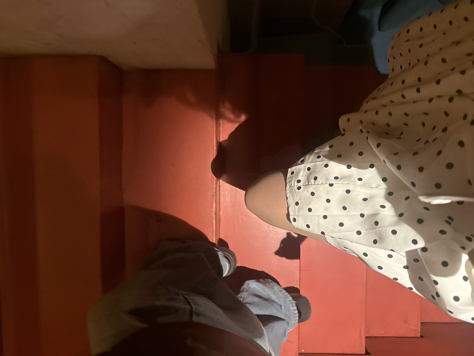 The photograph is taken from a high-angle perspective, looking down at a set of stairs. The stairs are a deep red or orange color. Two individuals are present in the image. One individual is wearing a light-colored dress with black polka dots, and another is wearing blue jeans. The feet of both individuals are visible on the stairs. The environment appears to be indoors. The photograph does not reveal any specific details about Yerevan, Armenia.