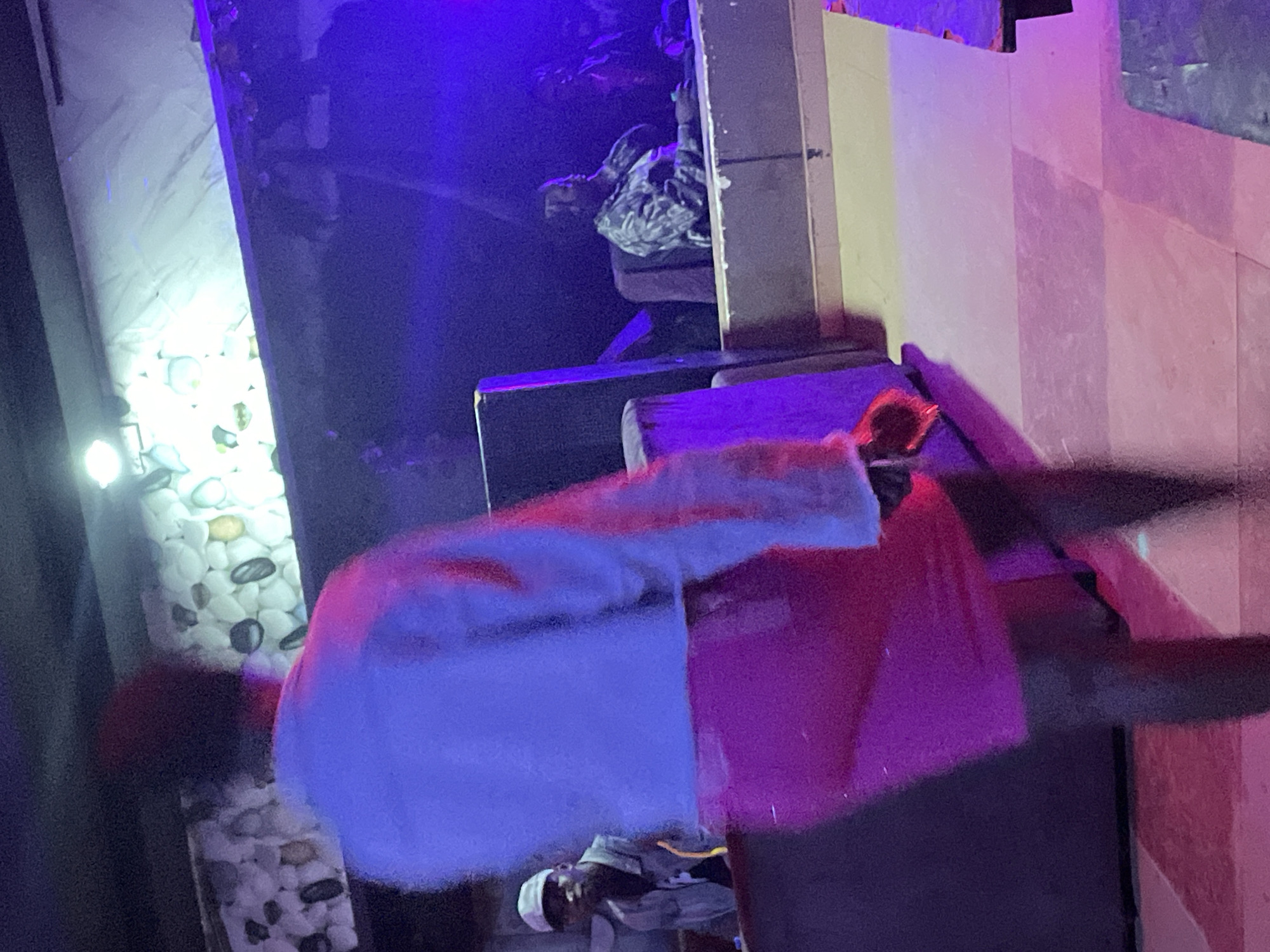 In Durumi, Nigeria, a dimly lit indoor space, possibly a club or event venue, is depicted. The scene is characterized by vibrant purple and pink lighting, reflecting off various surfaces. In the foreground, a person from behind is captured, wearing a white jacket over a pink skirt. Their form is slightly blurred due to motion or low light.
Further back, to the left, a person wearing a light-colored cap and a grey denim jacket is seated, looking towards the camera. To the right, another individual is seated in a chair, wearing a camouflage-patterned shirt. They are resting their hand on their chin, seemingly engaged in thought or conversation. The environment features a stone-like textured wall in the background, illuminated by bright lights, and a checkered floor with purple and pink hues. The overall atmosphere suggests a lively evening gathering.