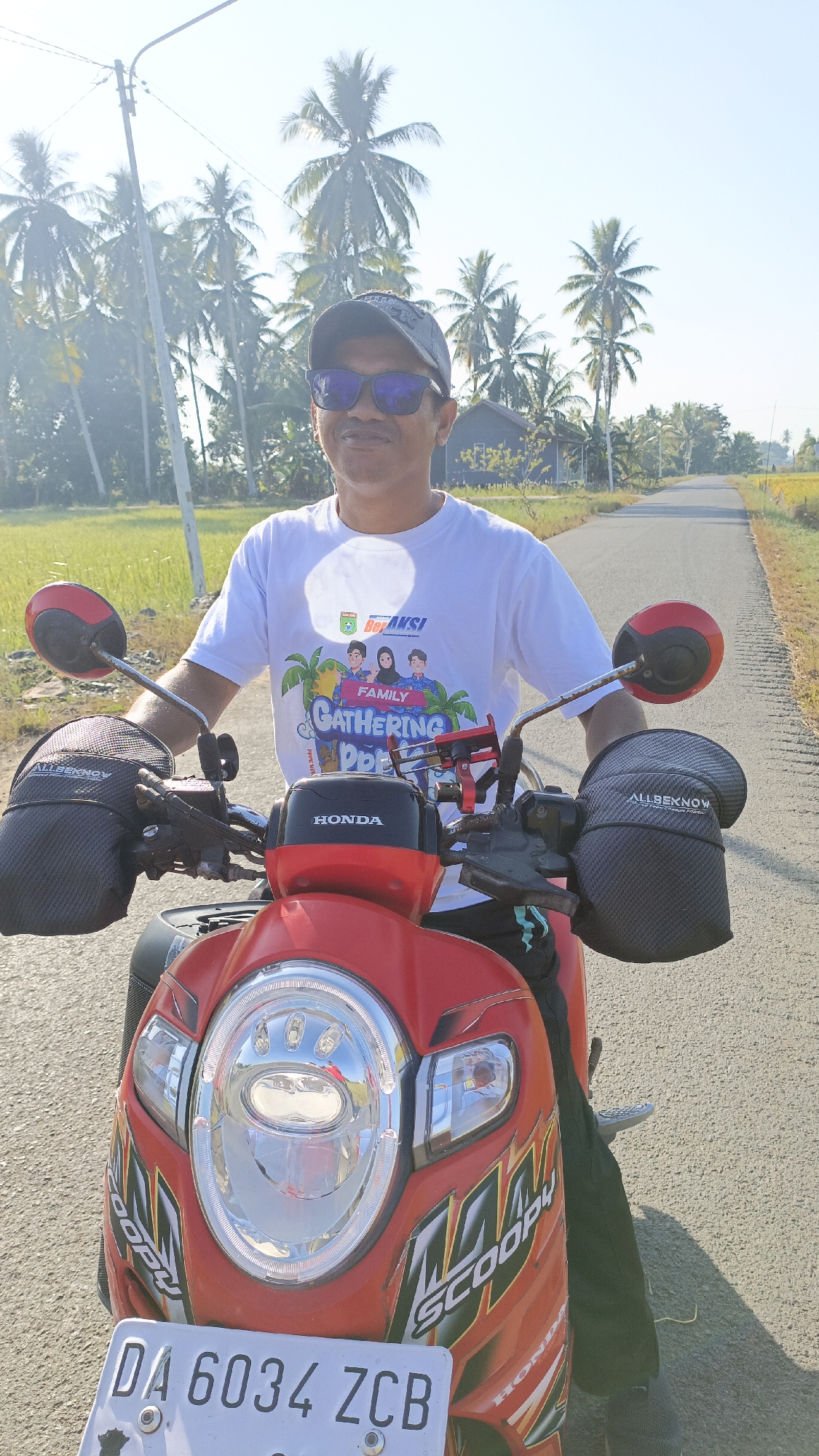 A man wearing a white t-shirt with a graphic design and a grey baseball cap is sitting on a red Honda Scoopy motorcycle on a paved road. He is wearing sunglasses and appears to be smiling. The motorcycle has "SCOOPY" written on the side. The background shows a rural landscape with palm trees and a clear blue sky. The license plate on the motorcycle reads "DA 6034 ZCB". The scene is set in Batarang, Indonesia, a region characterized by agricultural fields and abundant palm trees, suggesting a location outside of a dense urban center.