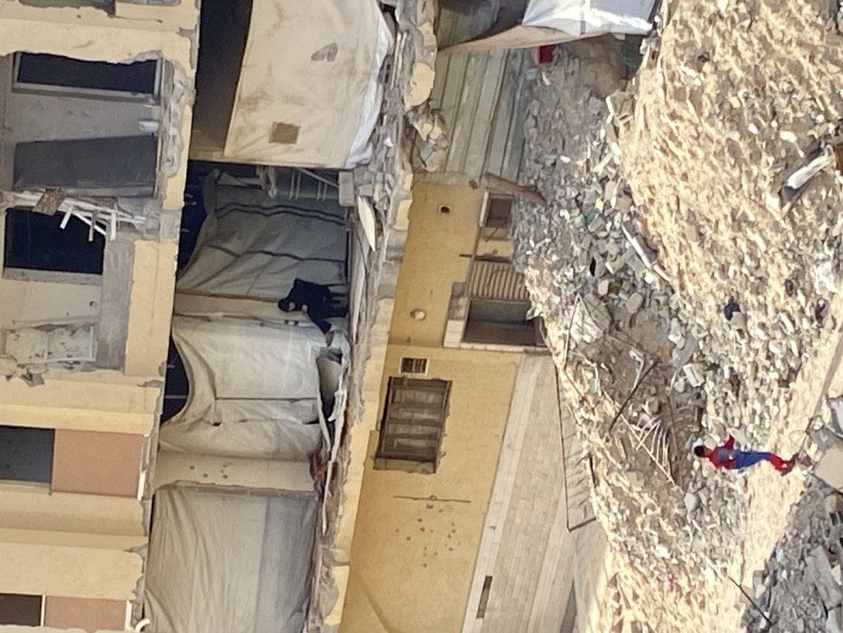 This image depicts a scene of extensive urban destruction in Khan Yunis, Palestinian Territory. The central focus is a severely damaged multi-story building with exposed structural elements and missing wall sections. On an intermediate floor of this damaged structure, an adult individual, dressed in dark clothing including a head covering, sits on a black chair within a makeshift enclosure constructed from white tarpaulins. This individual is observed leaning forward, appearing to engage with an object held in their lap. Directly below, a lower section of a building with a yellow facade exhibits multiple bullet impact points and features a wooden-framed window. The ground in the foreground is covered by large, irregular piles of pulverized concrete, rebar, and general construction debris. A young child, wearing a red and blue "Spider-Man" costume, is visible traversing this rubble, stepping on the uneven fragments. Another section of the damaged building in the upper right reveals additional exposed rooms, shattered windows, and debris, including a visible white bucket on a ledge.