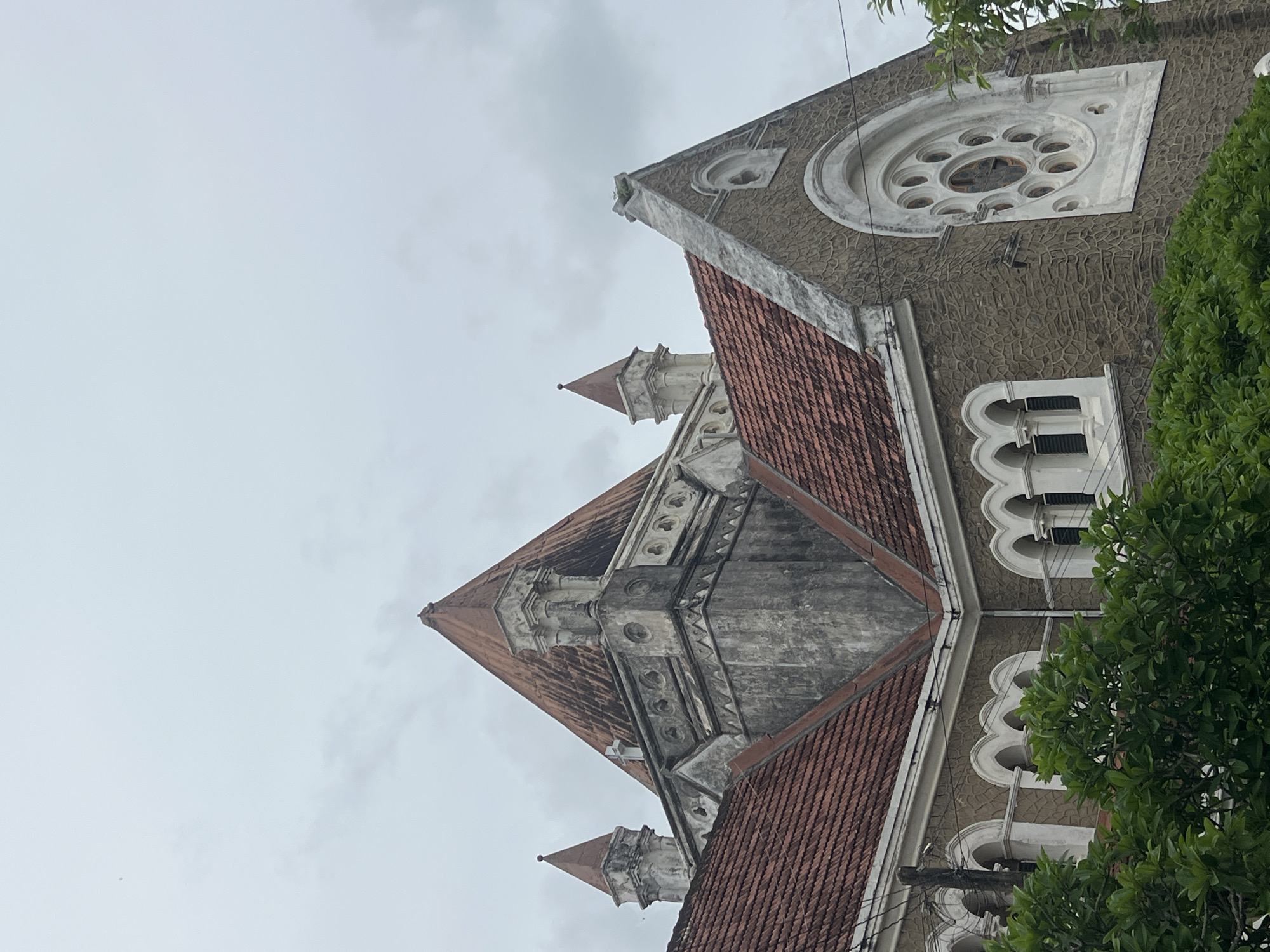 The image shows the exterior of a building with a large, conical, terracotta-tiled roof, typical of colonial architecture. The building has stone walls and arched windows. A circular, rose window is visible on the right side of the building. Green foliage is in the foreground, obscuring the lower part of the structure. The sky is overcast. This building is likely the Dutch Reformed Church in Galle, Sri Lanka, known for its historical significance and architectural style.