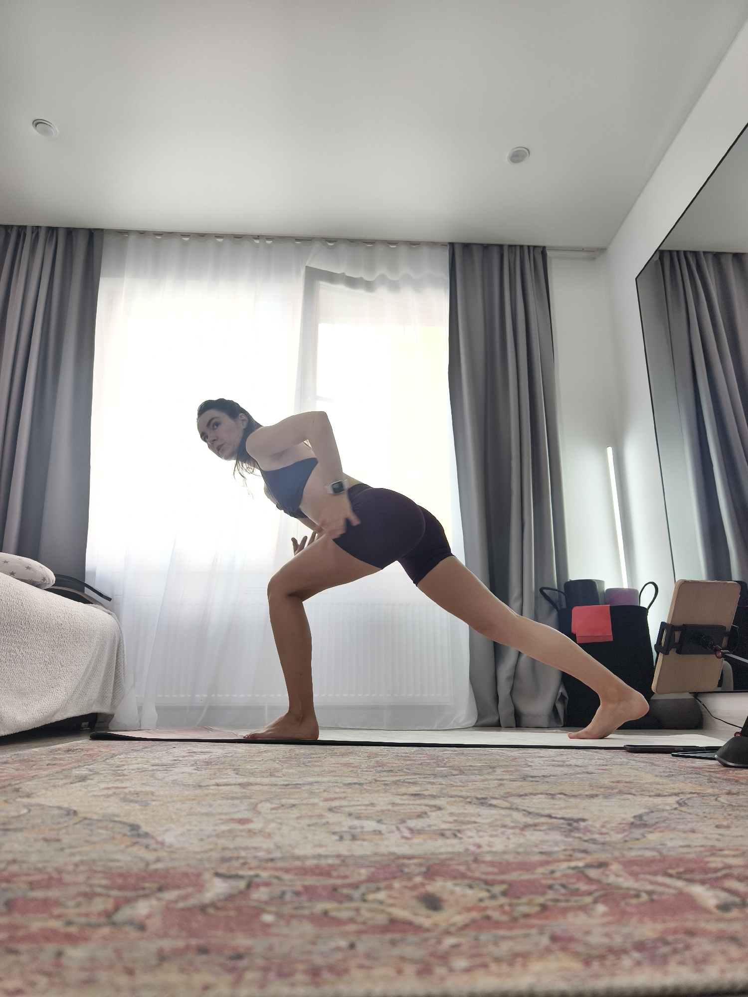 A woman with dark hair is performing a yoga pose in a brightly lit room, possibly a bedroom or studio. She is wearing a dark sports bra and dark shorts. Her right leg is extended back in a lunge position, and her left leg is bent. She is leaning forward, with her left arm extended towards the floor and her right hand on her hip. The room has light-colored walls and a large window with sheer white curtains and grey drapes. A rug with a patterned design covers the floor. To the right, there are several yoga blocks in a bag, a tablet on a stand, and a tall, thin lamp. To the left, a portion of a bed with white bedding is visible. The scene is an indoor residential setting. There is no information in the image to suggest it is located in Tyumen, Russia.