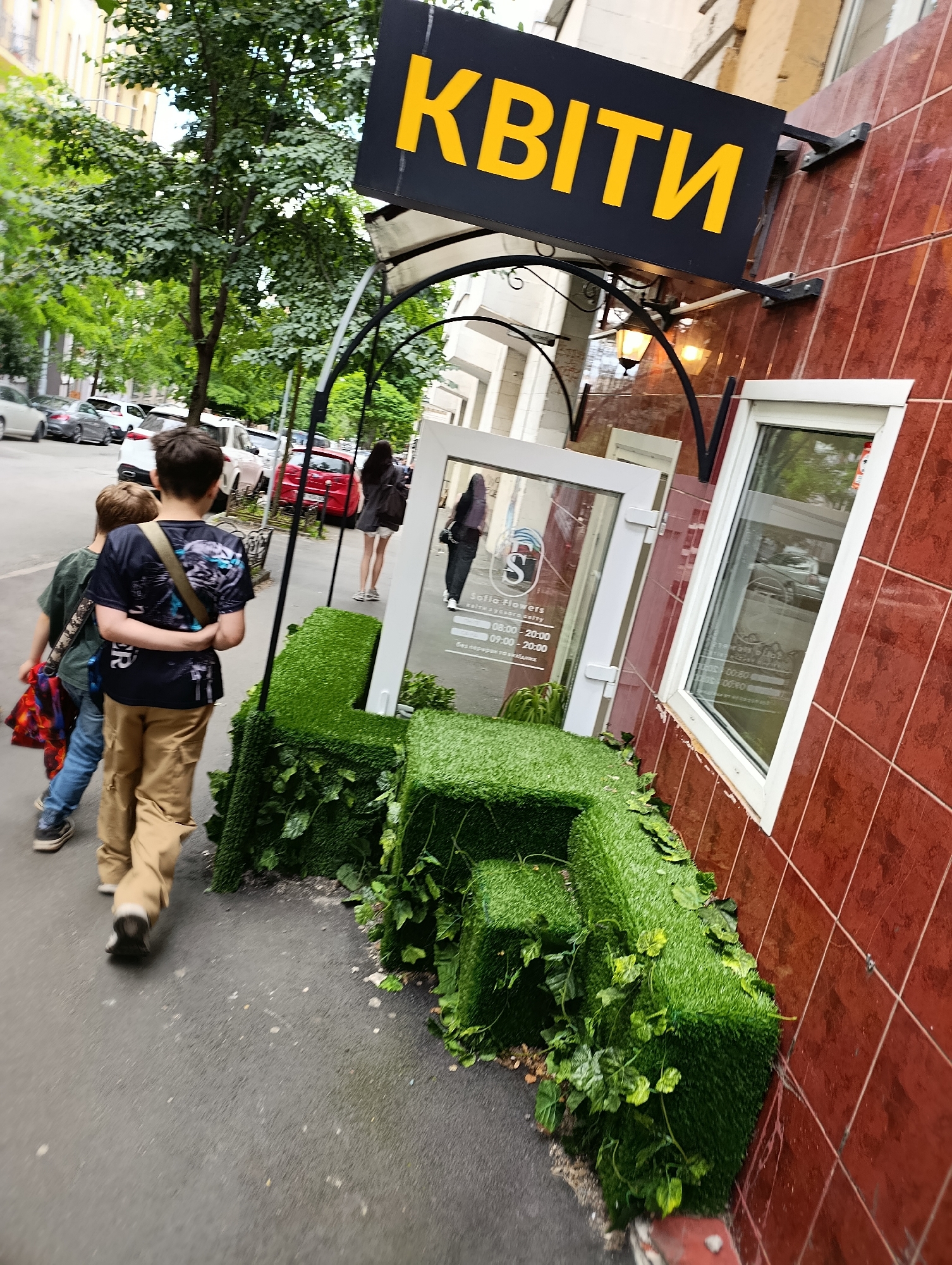 A low-angle, full outdoor shot shows a sidewalk scene in Kyiv, Ukraine. The dominant subject is a storefront with a large, yellow sign that reads "КВІТИ" (Flowers) in bold, sans-serif font. The sign is mounted on a black metal bracket attached to a red-tiled building. To the left of the building, a sidewalk runs alongside a street lined with parked cars, including a white SUV and a red sedan.
In the foreground, two boys are walking away from the camera down the sidewalk. The boy on the right, wearing a dark t-shirt and tan pants, has his arm around the shoulders of the boy on the left, who is wearing a green t-shirt and jeans and carrying a backpack with a colorful design. In front of the storefront, a decorative arrangement of green artificial turf cubes is placed along the sidewalk, intertwined with green vines.
Behind the boys, a woman in a black dress and white sneakers is walking away from the storefront. The storefront itself features a glass door and large glass windows. A sign inside the window displays text that includes "Sofia-Flowers" and operating hours. The overall environment suggests an urban setting on a cloudy day.