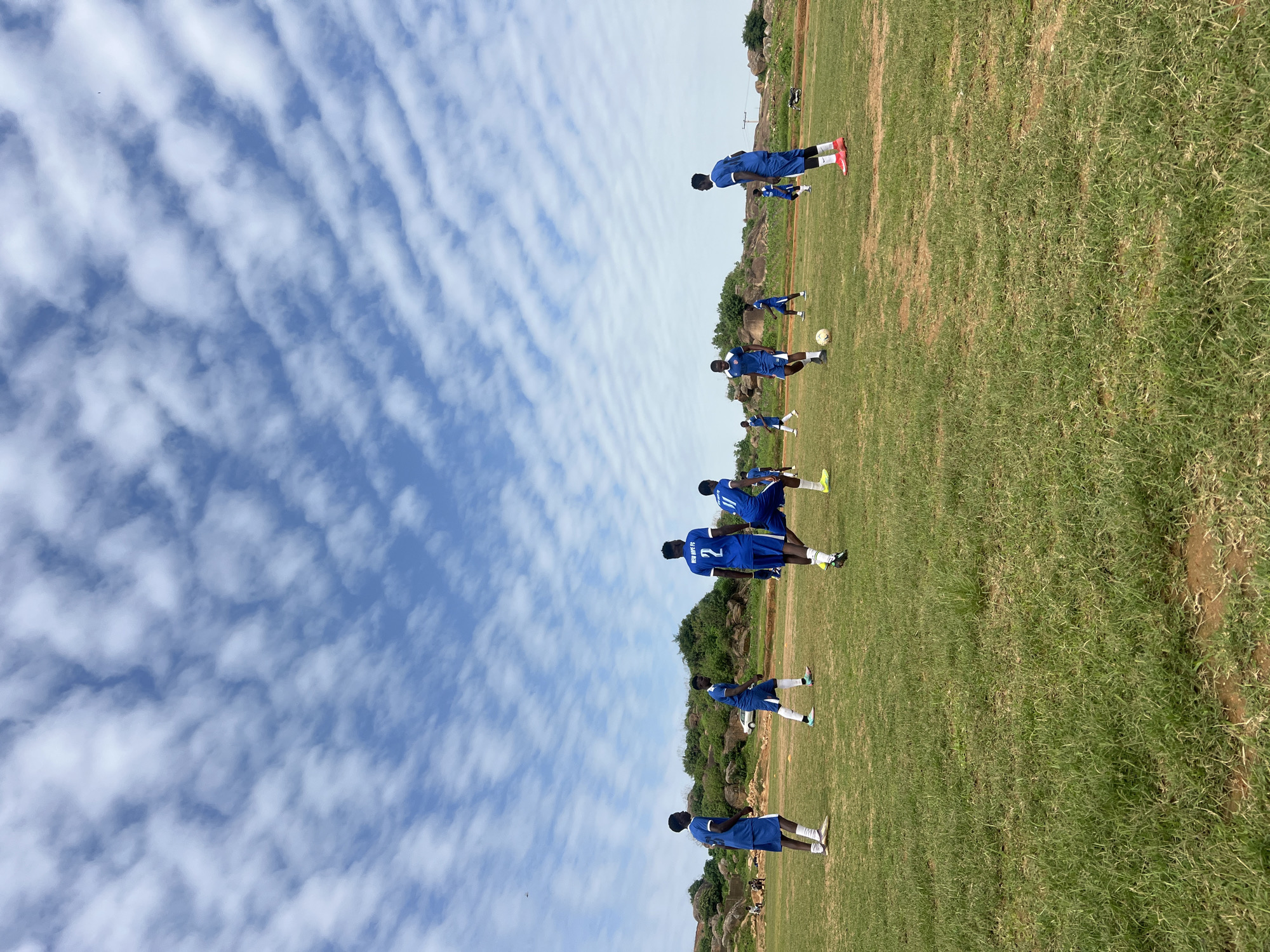 A group of young men, dressed in blue soccer uniforms with white trim, are engaged in a soccer practice on a grassy field under a partly cloudy sky. The field appears somewhat unkempt with patches of dry earth and sparse grass in some areas. In the background, there are rocky hills with sparse vegetation and a few trees. Several players are actively participating in the game, with one player in the foreground near the center of the frame dribbling a soccer ball. Other players are positioned around the field, some running, others standing. Two players in the mid-ground are facing each other, seemingly interacting during the game. In the far background, there is a white vehicle and some structures on the hills. The sky is a mix of blue and white clouds, with the clouds appearing somewhat striated. The overall impression is a casual soccer practice taking place in a somewhat rugged, outdoor environment. The inscription "NEW HOPE FC" is visible on the back of some of the players' jerseys. This scene is consistent with an outdoor sports facility in Jos, Nigeria, known for its rocky terrain and open spaces.
