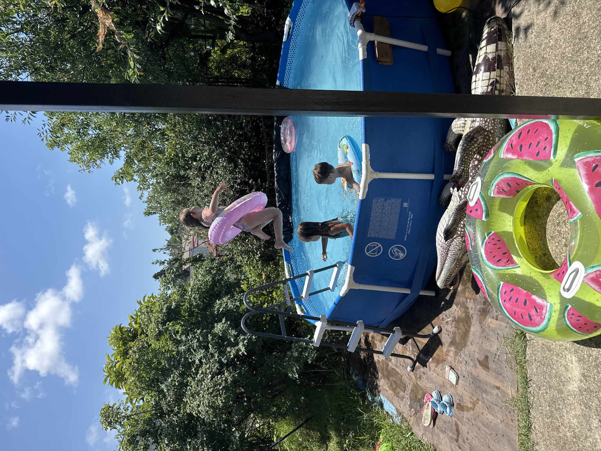 This is a vertical, top-down view of an outdoor scene on a sunny day, likely in Sochi, Russia, featuring a temporary above-ground pool filled with blue water. Two children are present in the pool, one wearing a blue swim ring and the other, a girl, is partially submerged. Another child, a girl, is in the process of jumping into the pool, her body mid-air, wearing a pink inflatable ring. In the foreground, a large, green inflatable ring decorated with watermelon slices lies on the ground next to a grey, checkered inflatable alligator. Scattered on the wet ground are a pair of pink flip-flops and a pair of blue Crocs. The pool is surrounded by lush green trees and foliage, with a clear blue sky and scattered white clouds visible above. A ladder is attached to the side of the pool, providing access. The lighting suggests it is daytime.