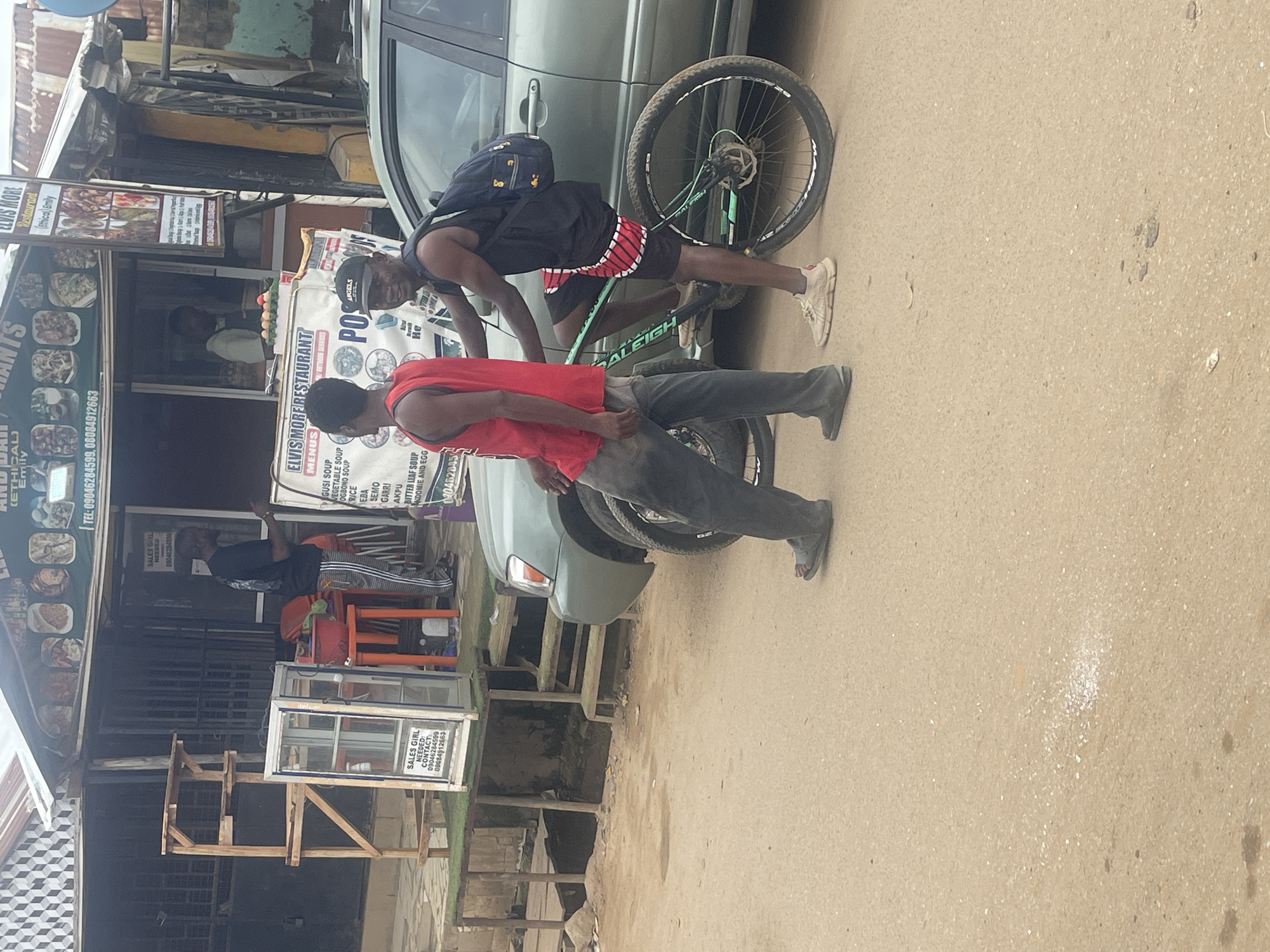 This is a vertical, eye-level, outdoor shot taken in Abuja, Nigeria. The scene depicts two men and a bicycle on a dusty road. The man on the left, wearing a black tank top and red and black shorts, is leaning over the bicycle, appearing to adjust it or hold it in place. He has a dark backpack slung over his shoulders. The man on the right, dressed in a red tank top and gray pants, is standing near the bicycle, his arm extended slightly forward.
Behind the men and bicycle, a light green car is parked. To the left of the car is a small roadside restaurant with various signs advertising food and services. One sign prominently displays "ELVIS MORE RESTAURANT" with a menu listed below, including items like "GUSI SOUP," "VEGETABLE SOUP," and "RICE." Another sign indicates "SALES GIRL NEEDED" with contact numbers. The overall impression is a busy, everyday scene at a local eatery. The lighting suggests it's daytime, likely late morning or early afternoon, with overcast or hazy conditions given the soft light and lack of strong shadows. The ground is a mix of asphalt and dirt, common in such urban settings.