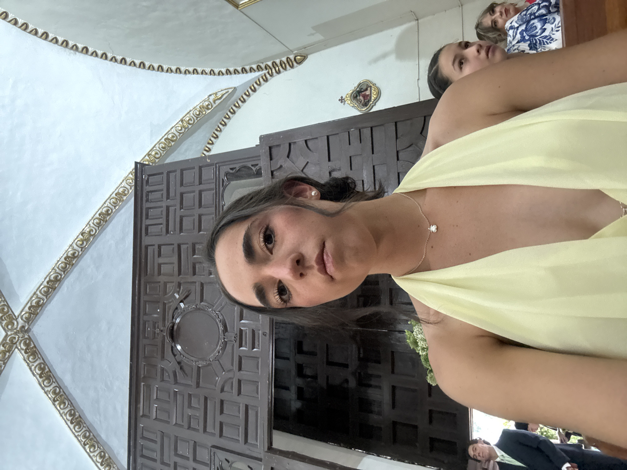 A medium shot shows a woman in a yellow dress indoors. In the background is an ornately carved wooden door and ceiling decorations. The location is likely a church in Orizaba, Mexico. Two other women can be seen behind the main subject, and a man in a suit is partially visible at the bottom.