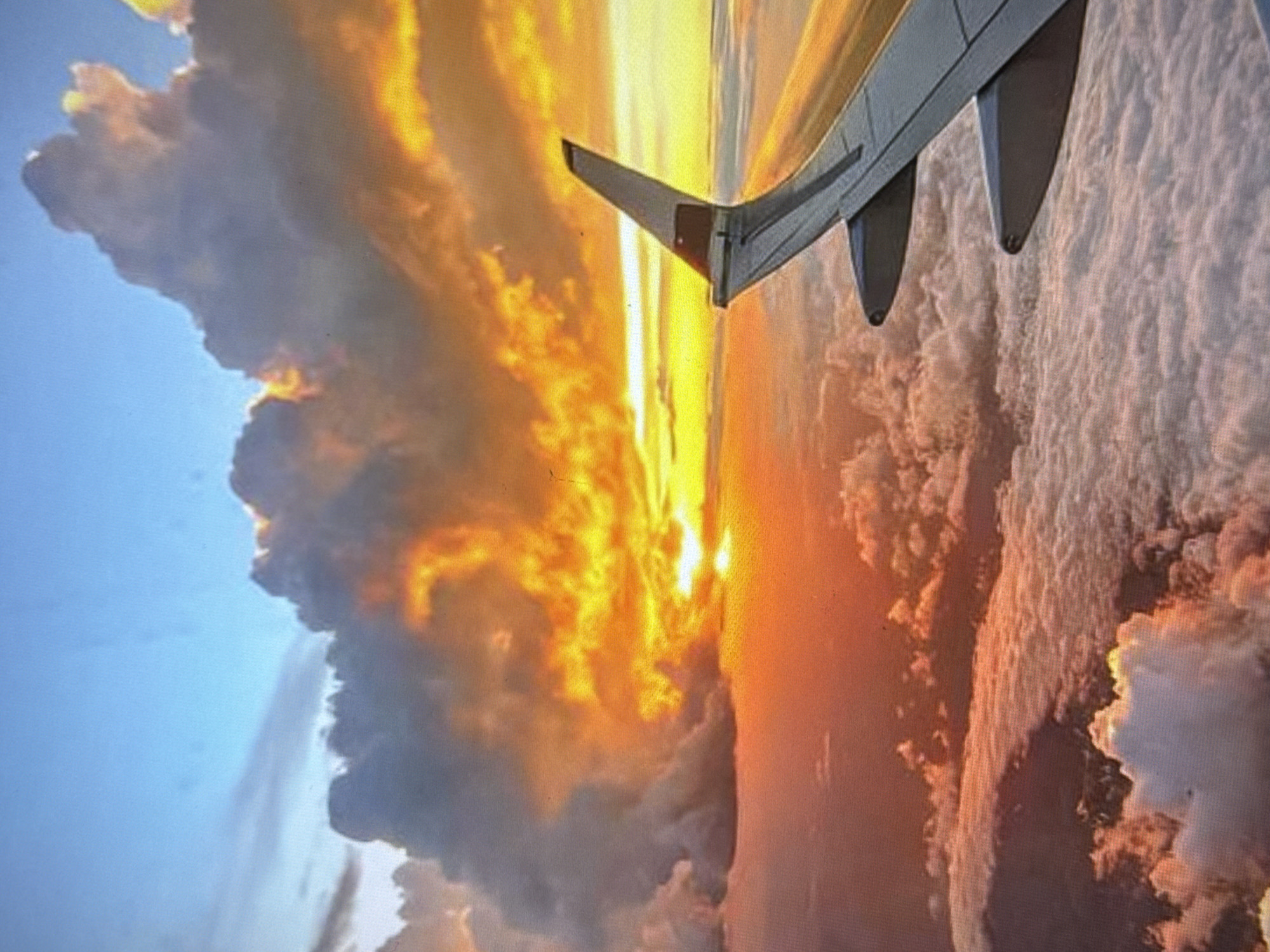 An aerial view captures a dramatic sunset from an airplane window. The sky is filled with vibrant oranges and yellows, illuminated by the setting sun, which is partially obscured by thick, billowy clouds. The clouds themselves are a mix of dark grays and brilliant oranges, creating a fiery spectacle. Below the clouds, a sea of white clouds stretches across the horizon. In the foreground, the wing of an airplane is visible, with its wingtip pointing towards the right side of the frame. The image does not depict any specific location in Miass, Russia.