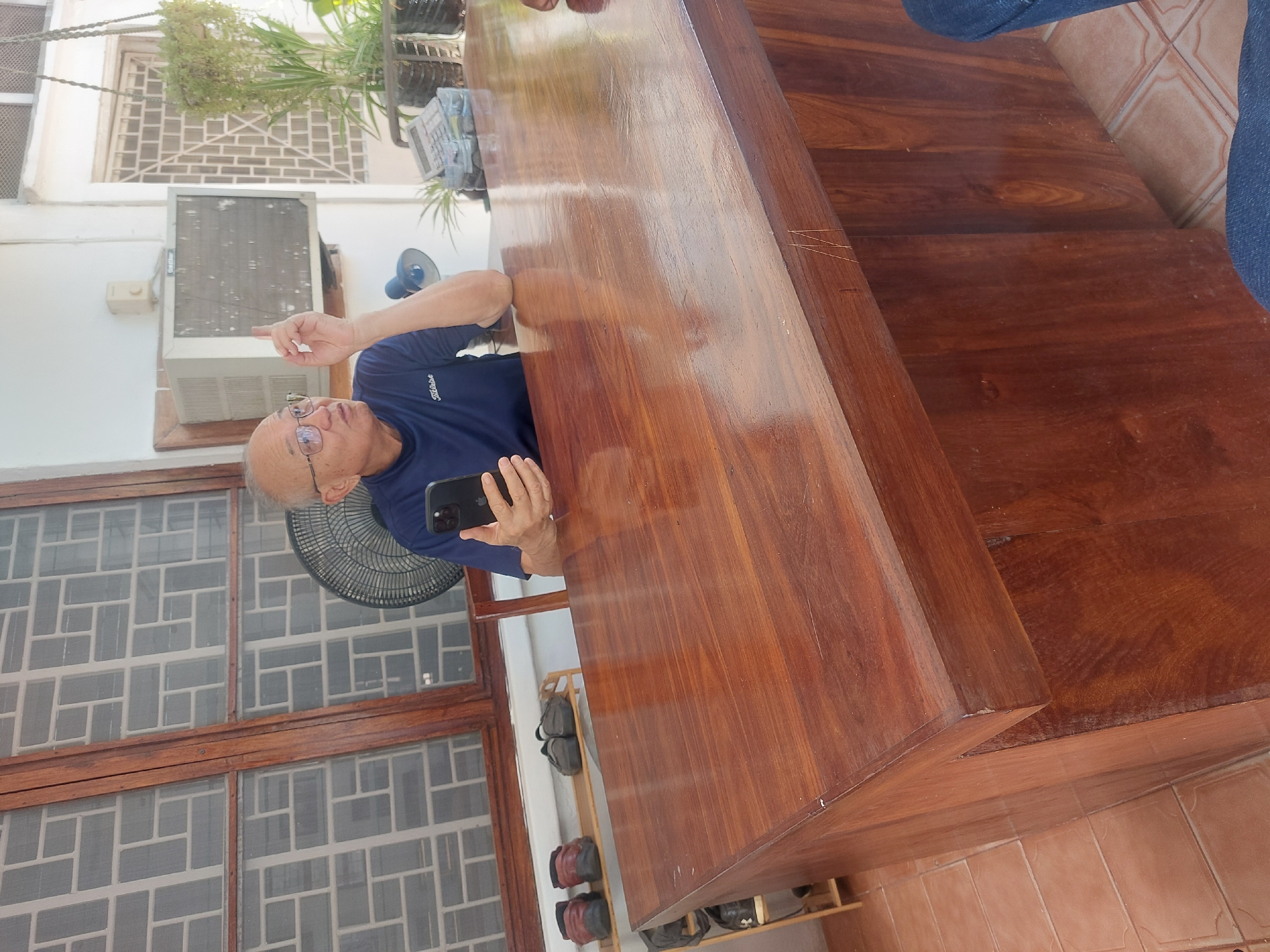 The image depicts an indoor or semi-outdoor area, likely a covered patio or large open room, in what is identified as Dar es-Salaam, Tanzania. The scene is brightly lit, suggesting daytime.
The primary subject is a man, appearing middle-aged or older, positioned on the left side of a long, highly polished dark wooden table or counter. He wears glasses and a dark blue polo shirt with the brand name "Titleist" visible on the chest. He holds a smartphone, possibly an iPhone given the camera array, in his left hand and is actively pointing with his right index finger, indicating something out of frame. His expression is neutral and engaged, suggesting he is explaining or drawing attention to something.
Behind the man, a small black fan is visible, and further back on a white wall, a wall-mounted air conditioning unit is present, with some unreadable white text on its casing. The background features large windows with decorative white grates, allowing ambient light into the space. Several potted and hanging plants are also visible, adding to the tropical feel. Near the floor, a wooden shoe rack holds several pairs of dark-colored sandals or slippers. The floor is tiled with light brown or orange square tiles. A second person's lower leg, wearing blue jeans, is partially visible in the upper right corner, standing near the end of the wooden table. The large wooden table or counter, extending across the image, has a smooth, glossy finish that reflects light. The overall setting implies a comfortable environment in a warm climate.