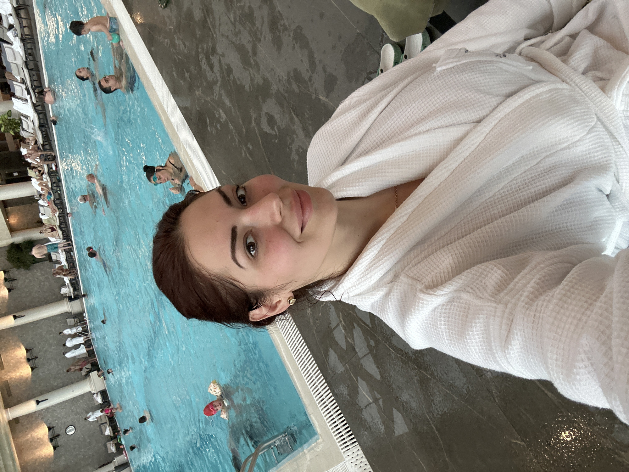 The image shows a woman in a white bathrobe taking a selfie next to a large indoor swimming pool. The pool is populated with several people swimming and relaxing. On the side of the pool opposite the woman, there are lounge chairs and tables, some of which are occupied. The floor surrounding the pool is gray and appears to be wet. The location is likely a spa or resort with indoor pool facilities. No information indicates the location is Kazanlak, Bulgaria.