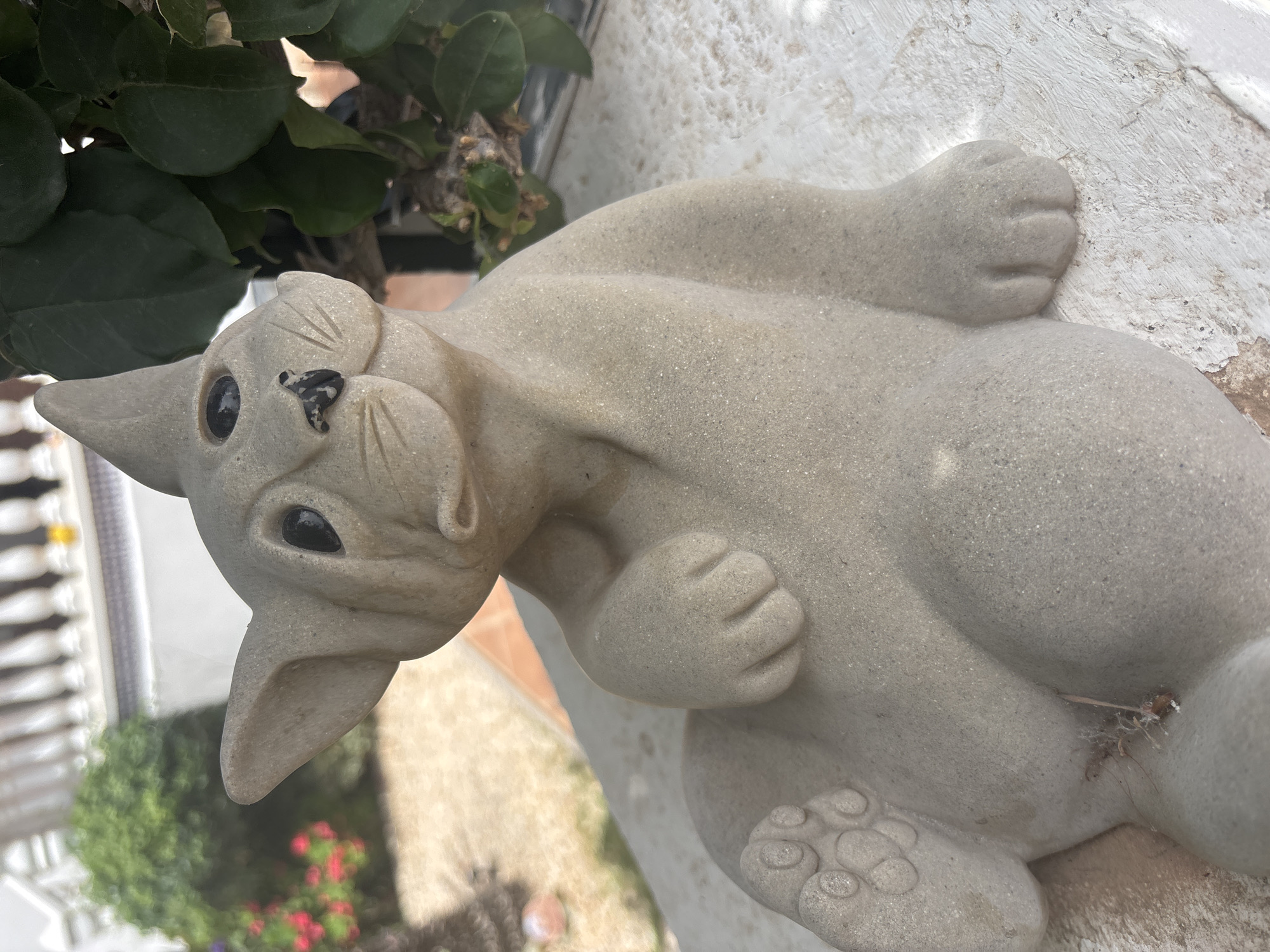 A light gray, stone-like statue of a cat is positioned outdoors, resting on a white, textured surface. The cat statue is in a reclining pose, with its head tilted upwards and to its left, revealing a simplified facial structure with black oval eyes and a small black nose. Its left paw is raised, resting near its face, and its right paw is extended forward. The background shows blurred greenery and what appears to be a building with white railings. The scene suggests an outdoor patio or garden setting.