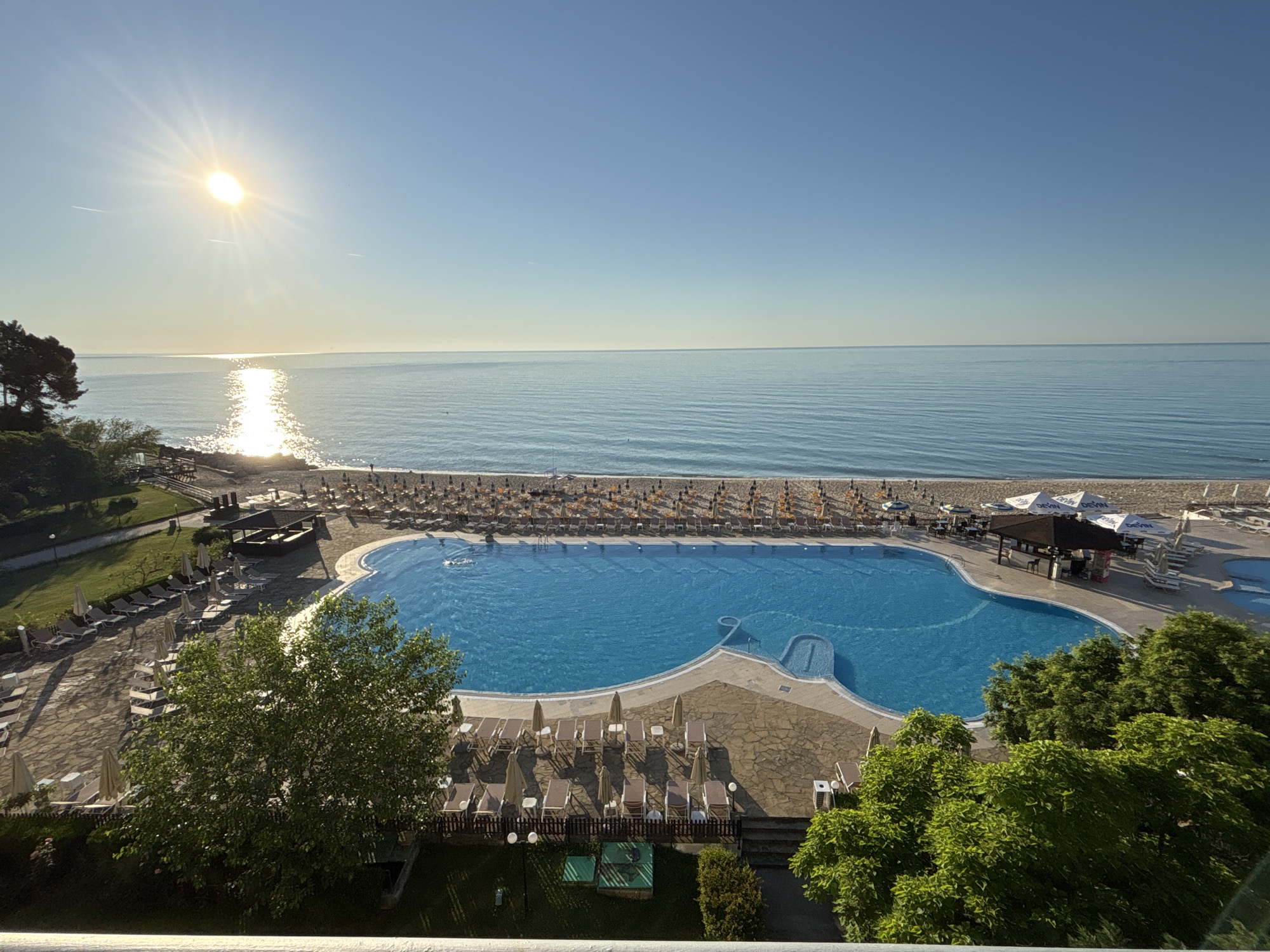 The image captures a resort scene in Varna, Bulgaria, during a bright, sunny morning. The foreground features lush green trees and a meticulously maintained outdoor swimming pool with clear blue water. Lounge chairs and umbrellas are arranged around the pool, some occupied by sunbathers. Beyond the pool, a sandy beach is lined with rows of umbrellas and lounge chairs, extending towards the calm, blue sea. The sun shines brightly in the clear sky, casting a shimmering reflection on the water. The overall impression is one of a serene and inviting vacation destination.