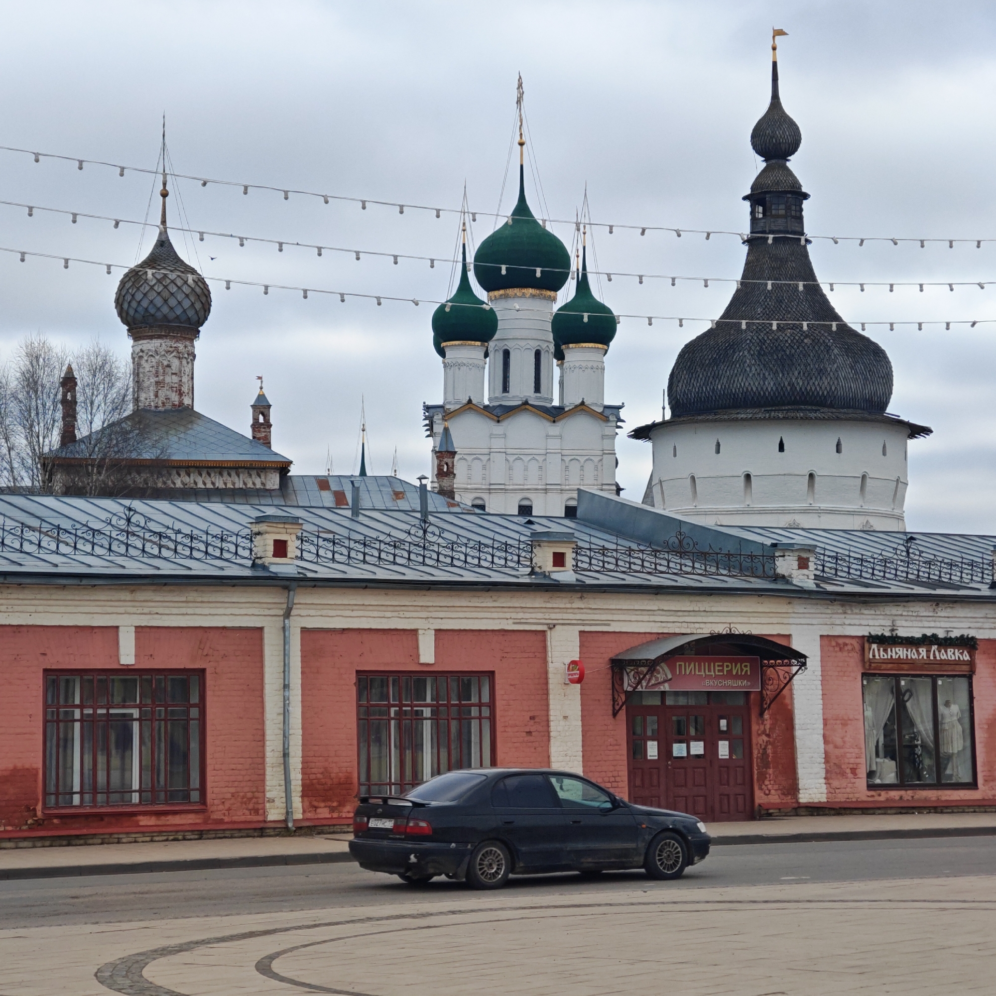 The image shows a street scene in Rostov, Russia, featuring several architectural landmarks. In the foreground is a red brick building with a gray roof and windows with dark frames. Signage is visible above the entrance for "ПИЦЦЕРИЯ (ВКУСНЯШКИ)" and "ЛЬНЯНАЯ ЛАВКА." A black car is parked or driving on the street in front of the building. Behind the brick building, there are three prominent towers with distinctive tops: one has a gray dome, another with three green domes, and a taller tower with a black, layered, wooden top. Overhead, strings of lights are suspended. The sky is overcast. The street is paved with patterned stones. There are trees in the background. The image conveys an urban environment with a mix of commercial activity and historical architecture.
