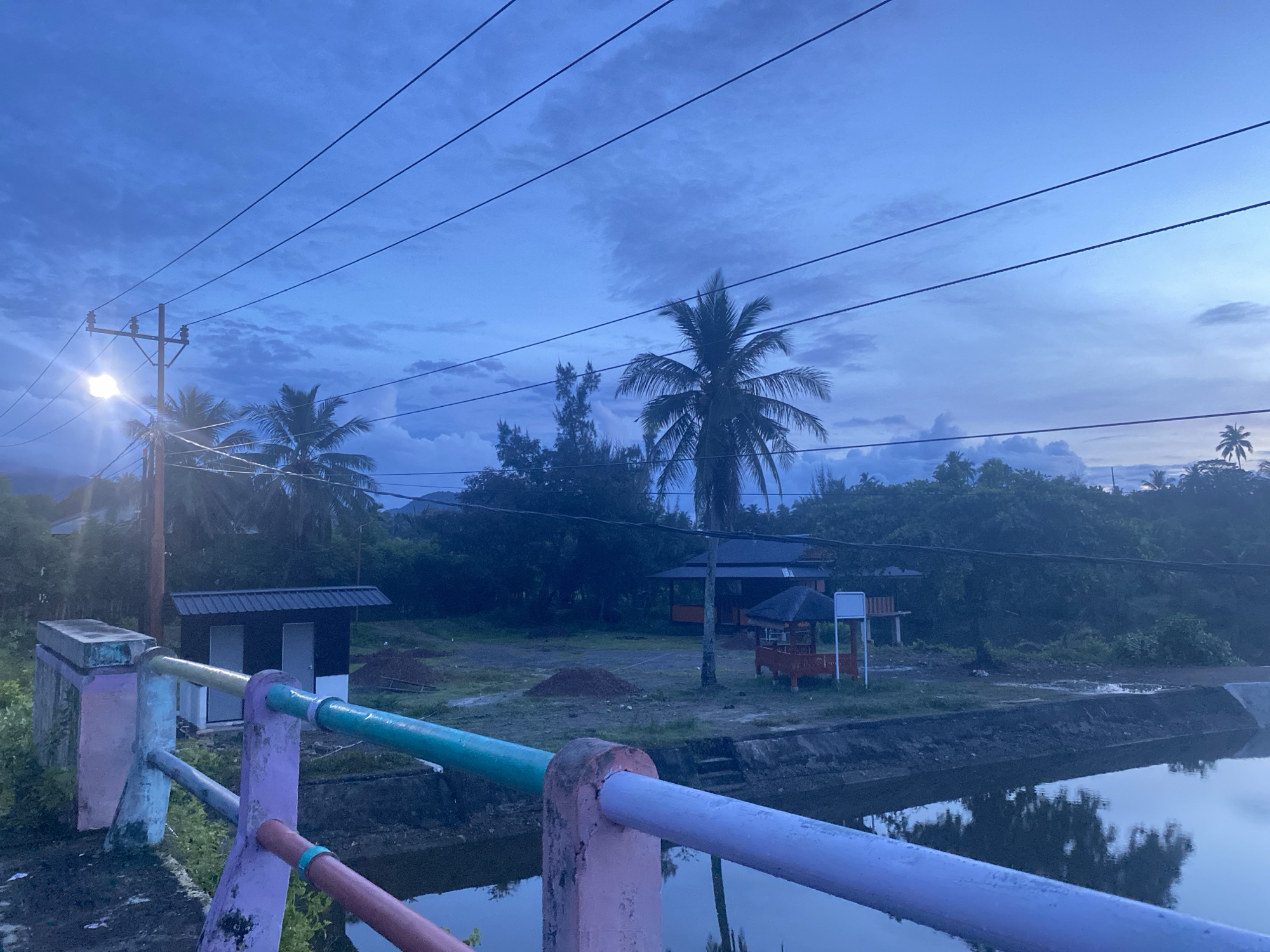 The image depicts a serene twilight scene in Aneuk Paya, Indonesia. A bridge railing, painted in vibrant pastel colors of pink, purple, and teal, dominates the foreground. Beyond the bridge, a tranquil body of water reflects the dusky sky. The midground features a small, dark-colored building with a metal roof and two white doors, flanked by lush green vegetation. Further back, several palm trees and other tropical foliage create a dense backdrop, interspersed with a few structures, including a red gazebo with a dark roof and an orange-hued building with a dark roof. Power lines stretch across the sky, one pole illuminated by a bright light. The sky itself is a canvas of deep blues and grays, with scattered clouds hinting at the fading daylight. The overall atmosphere is peaceful and rural, characteristic of a quiet evening in this Indonesian locality.