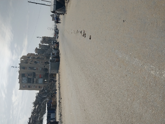 The image depicts a scene of extensive urban destruction in the city of Khan Yunis, Palestinian Territory, under an overcast sky during daytime. The foreground is dominated by a wide, unpaved or heavily worn asphalt road surface, showing scattered debris and small indentations.
On the left side of the midground, a massive pile of grey rubble from a collapsed structure rises significantly. Adjacent to this rubble, at its base, are several small, makeshift market stalls or shelters, one with a blue awning, and a wooden table displaying items. A person in dark attire is partially visible near these structures.
Centrally, a severely damaged multi-story residential building, approximately seven to eight stories tall, stands partially intact but with significant structural compromise, missing sections, and exposed interior elements. Its facade is light-colored, and large, dark letters, possibly graffiti, are visible on an upper floor. Antennas or communication equipment are mounted on its roof. In front of this building, on the roadside, a small, grey, rectangular kiosk or utility box is present. Another individual in dark clothing is visible near this kiosk.
The road extends into the background, flanked by more damaged structures and debris. On the right side of the road, several cars are visible, along with multiple pedestrians walking. Further right, makeshift tents or shelters are erected. Utility poles with overhead lines are discernible along the roadside. The overall environment indicates widespread architectural devastation, with ongoing human activity amidst the ruins.