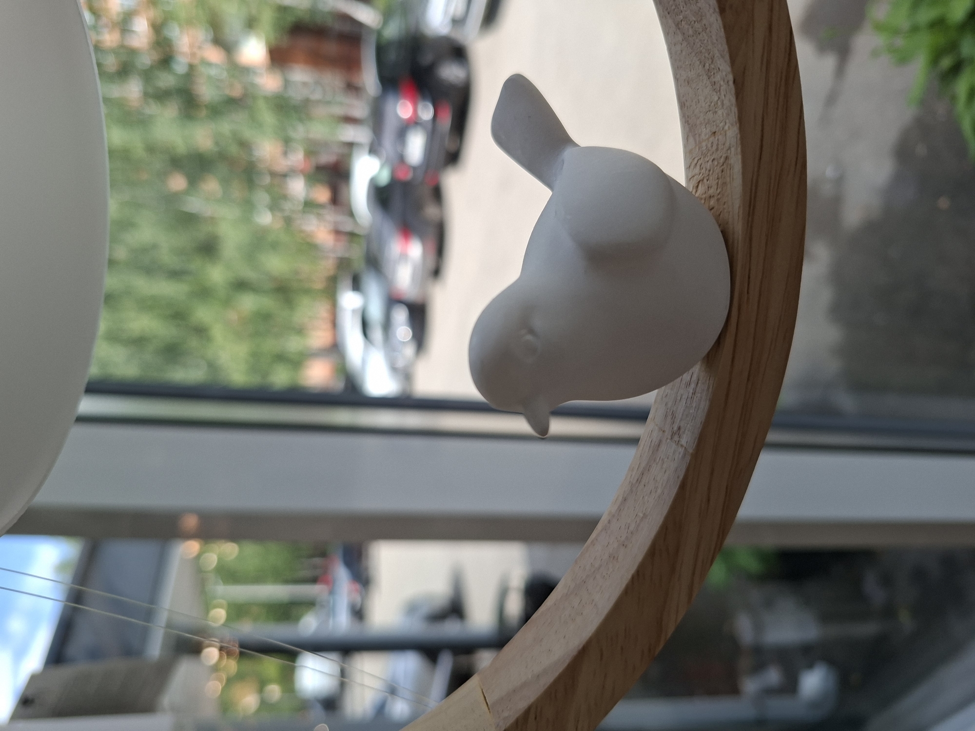 A white ceramic bird figurine rests on a curved wooden surface, positioned in front of a window that offers a blurred view of an urban environment. Outside, parked cars and trees are visible, suggesting a street or parking lot scene. The lighting indicates it is daytime, with sunlight filtering through the trees and reflecting on the window. The overall impression is one of a decorative object placed in a domestic or commercial setting, overlooking an outdoor space. It is not possible to determine the specific location within Izhevsk, Russia, based on the visual information provided.