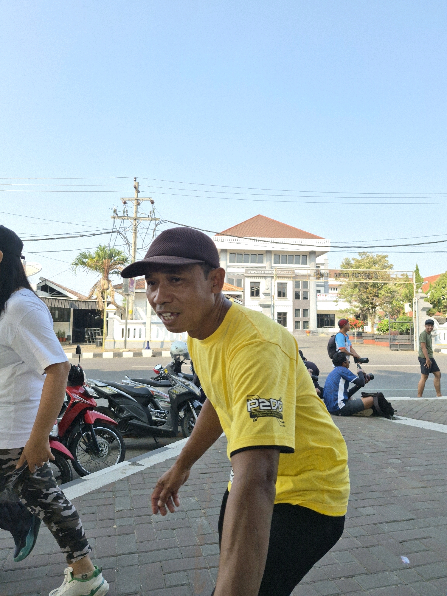 In the city of Purworejo, Indonesia, a man wearing a dark brown baseball cap and a yellow t-shirt with "P2DD" printed on it is in the foreground, looking towards the camera with his mouth open. He is standing on a brick-patterned sidewalk. To his left, a person wearing a white t-shirt and camouflage pants is walking away from the camera. Behind them, several motorcycles are parked along the street. In the background, a large building with a brown roof and white walls, identified by a sign as "SPKAD KABUPATEN PURWOREJO" and "KANTOR BUPATI PURWOREJO," stands under a clear blue sky. Other individuals are visible in the mid-ground, some holding cameras. The scene suggests a public gathering or event is taking place outdoors.