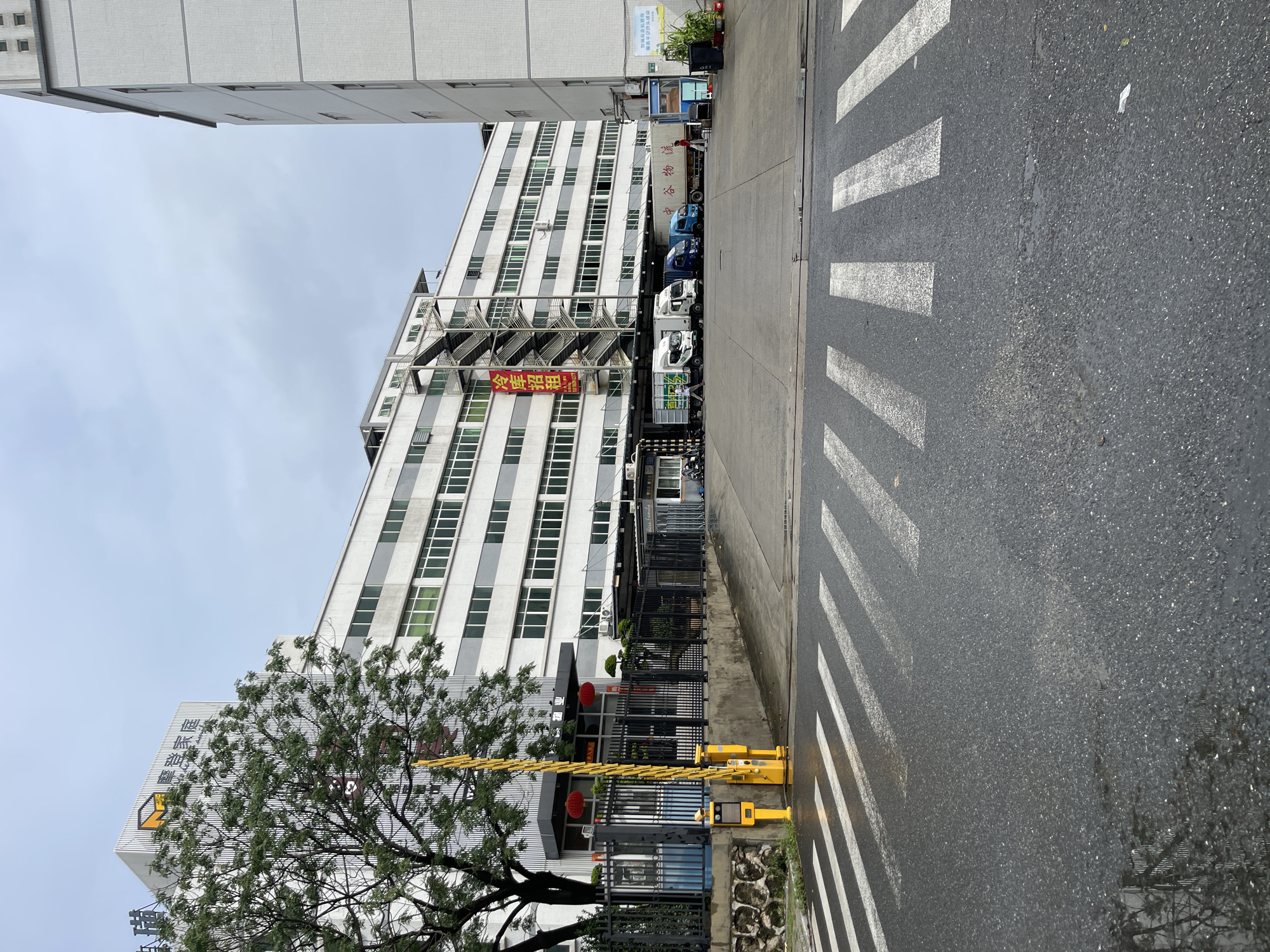 The image shows the exterior of a large, multi-story industrial building in Panyu District, China. The building is primarily white with gray horizontal accents and numerous windows. A large tree with green foliage is visible to the left of the building. In front of the building is a paved area with a pedestrian crossing marked on the road. A yellow gate arm is extended across a driveway, and a small booth is visible behind it. Several delivery trucks, including a refrigerated truck, are parked in the paved area in front of the building. A red banner with Chinese characters is hanging on the side of the building. The sky is overcast.
