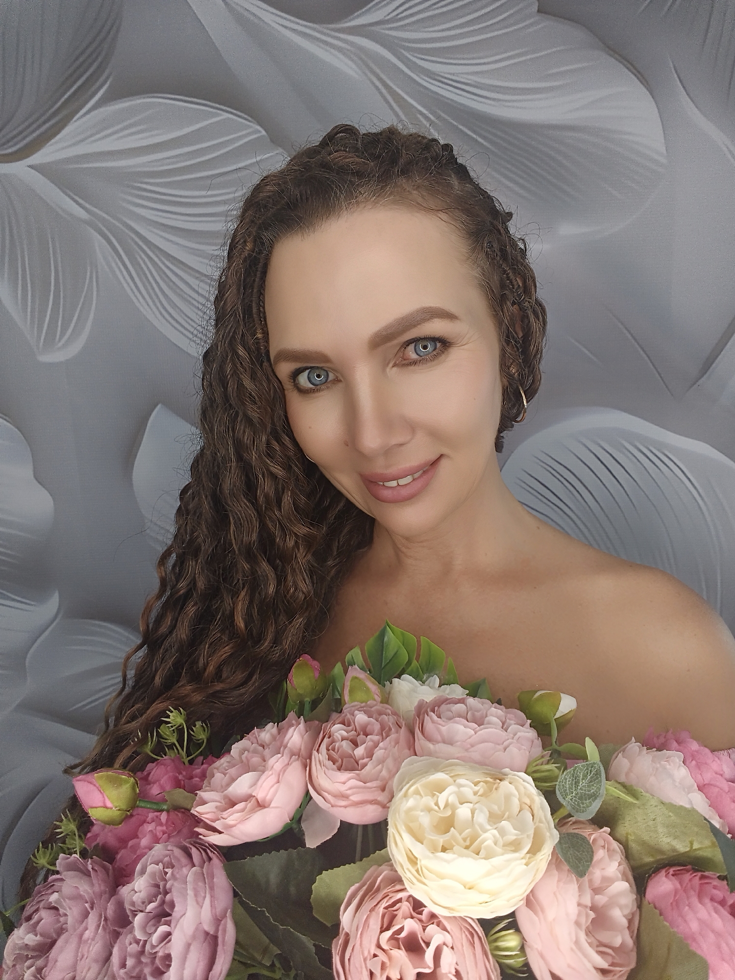 A medium shot of a woman with curly brown hair, blue eyes, and fair skin is shown from the chest up, holding a bouquet of pink and cream flowers. She is looking directly at the camera with a slight smile. She is bare-shouldered and wearing a gold hoop earring in her right ear. The background is a gray textured wall with a floral pattern. The lighting is soft and even.