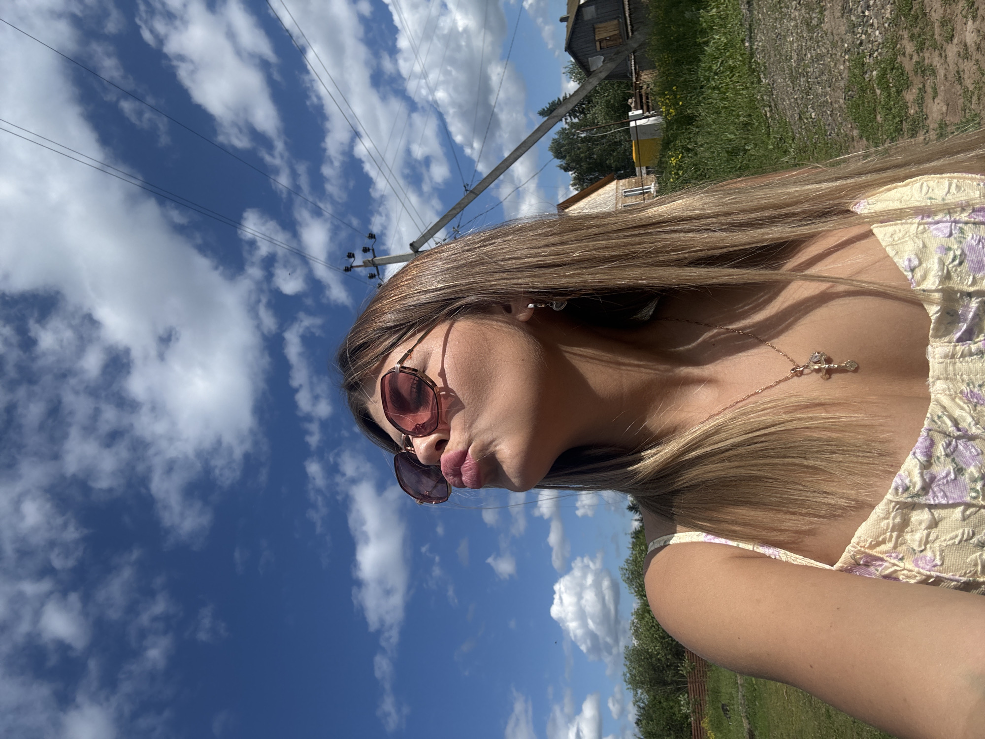 A young woman with long blonde hair and sunglasses is outdoors in what appears to be a rural area. She is wearing a light-colored, textured top with floral embroidery and a gold necklace with a cross pendant. Her lips are pursed as if blowing a kiss, and she is looking up towards a bright blue sky filled with white, fluffy clouds. In the background, there are trees, a few rustic buildings, and utility poles with wires, suggesting a village or countryside setting. The ground is covered in grass and gravel. The overall scene suggests a casual, sunny day.