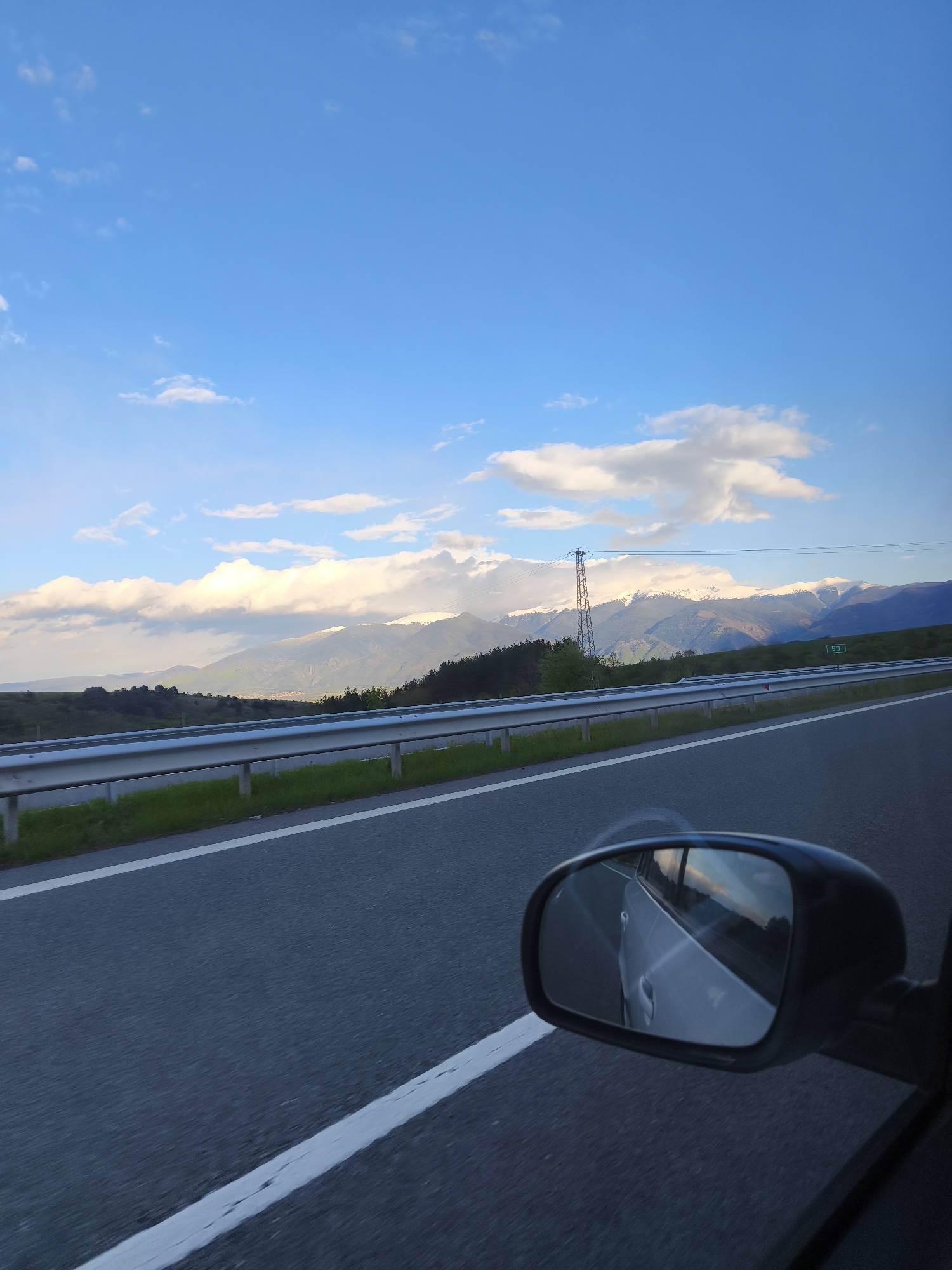 The image depicts a view from inside a moving vehicle, likely a car, traveling on a highway near Blatino, Bulgaria. The road is asphalt and has white lane markings. A metal guardrail separates the road from a grassy area. In the background, there are hills and mountains with snow-covered peaks, partially obscured by clouds. An electrical power transmission tower is visible in the middle distance. The sky is blue with scattered white clouds. The car's side mirror is visible in the foreground, showing a reflection of the car's rear side and the scenery behind it. A green sign with the number "53" is present near the guardrail.