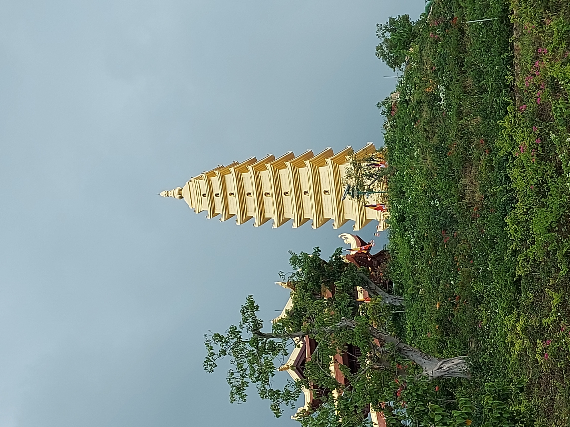 A tall, golden pagoda dominates the center-right of the frame, its multi-tiered structure reaching towards a pale, overcast sky. To the left of the pagoda, a traditional Vietnamese temple with a dark red roof and ornate golden trim is partially visible, obscured by lush green foliage. The foreground is covered in dense, varied greenery, including trees with visible branches and a profusion of smaller bushes and flowering plants in shades of green, pink, and white. Flags are visible on the temple grounds. The scene is a view of a temple complex on a hillside in Diên Khánh, Vietnam.