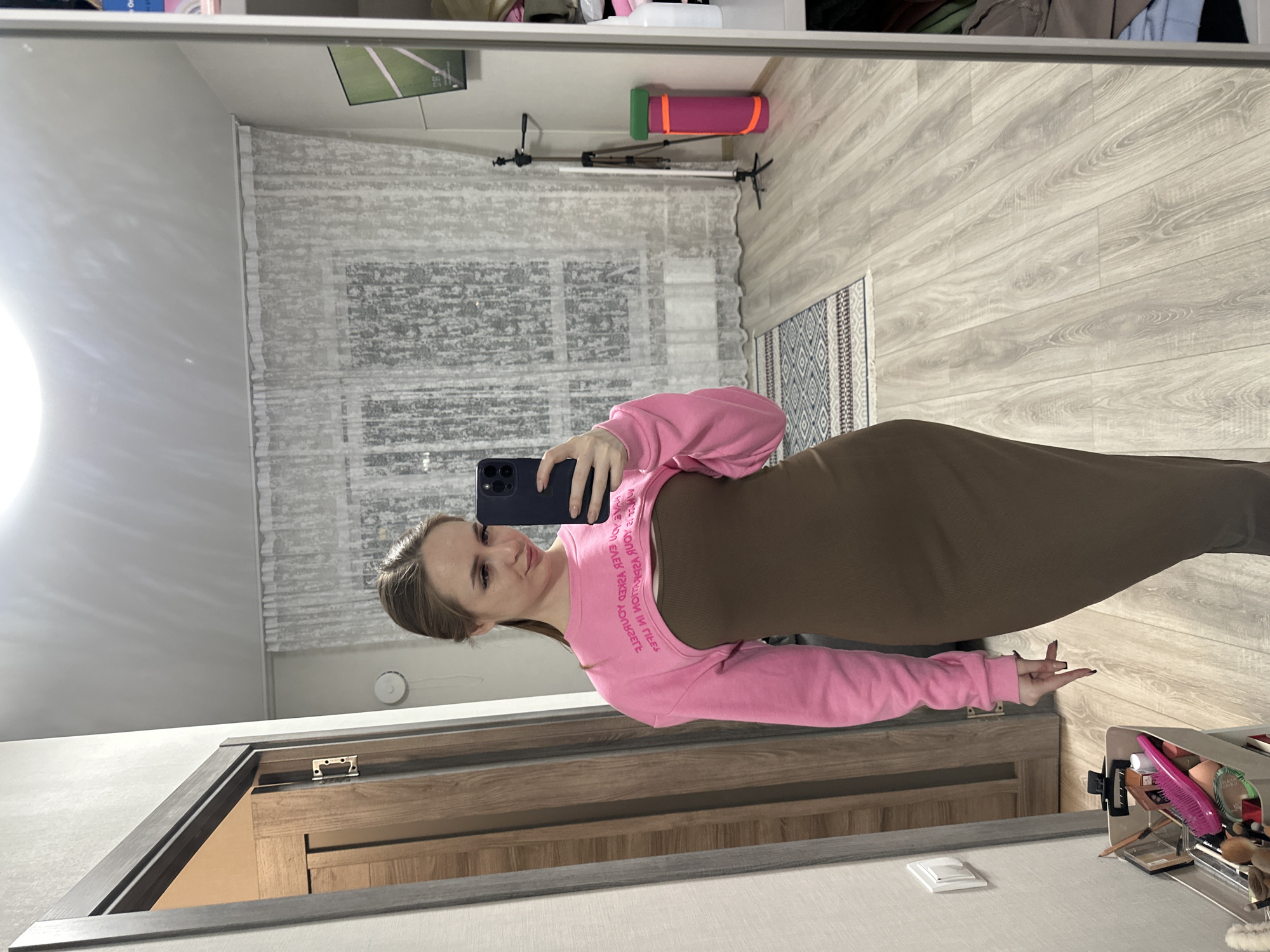 A young woman is captured in a mirror selfie, standing in what appears to be an indoor room. She is wearing a dark brown or olive-green form-fitting dress or long skirt, paired with a bright pink cropped long-sleeve top. The top features mirrored white text across the front, which reads "YOUNG IS YOUR BEAUTY, BEAUTY IS YOUNG". She holds a dark blue or black smartphone with multiple cameras, using it to take the picture. Her expression is a slight, pleasant smile, and her hair is pulled back.
The room visible in the reflection features light-colored wooden flooring, partially covered by a patterned rug with white and blue designs. A window with light grey, textured curtains is visible in the background, allowing for ambient light. Other objects reflected in the mirror include a pink and green yoga mat, a black tripod stand, and a flat-screen monitor mounted high on a wall, displaying what looks like a green sports field. Near the bottom right of the reflection, a vanity organizer holds various beauty items such as brushes and a pink comb. A wooden door frame with a visible hinge and a white light switch are also reflected. The setting appears to be a casual home environment, likely a bedroom or dressing area, during the daytime.