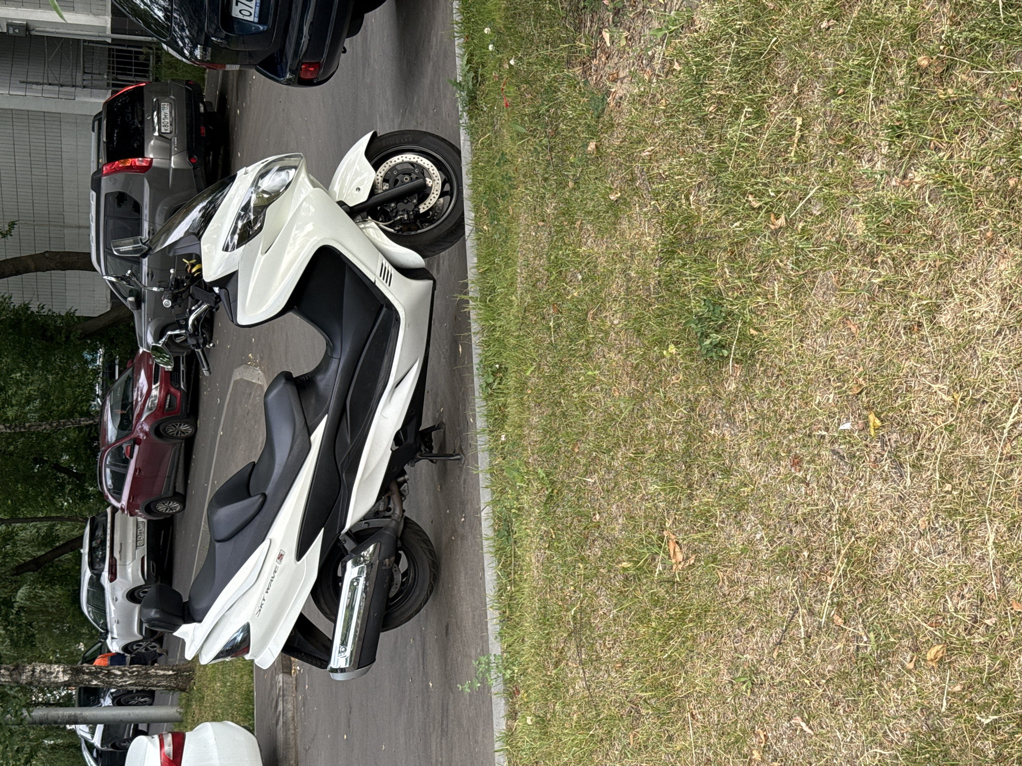 A white Suzuki Burgman 650 scooter is parked on an asphalt surface next to a grassy area. The scooter is oriented diagonally with its front end facing left. It has a black seat and a black windscreen. Several cars are parked in the background. To the left of the scooter, a white car is partially visible. Behind the scooter, a red Kia Rio sedan is parked, and further back, a gray Nissan X-Trail SUV and a dark-colored Hyundai Solaris sedan are visible. The environment appears to be a residential area in Moscow, Russia, as indicated by the license plate format on the Nissan X-Trail which reads "K801MV 199". The weather is overcast.