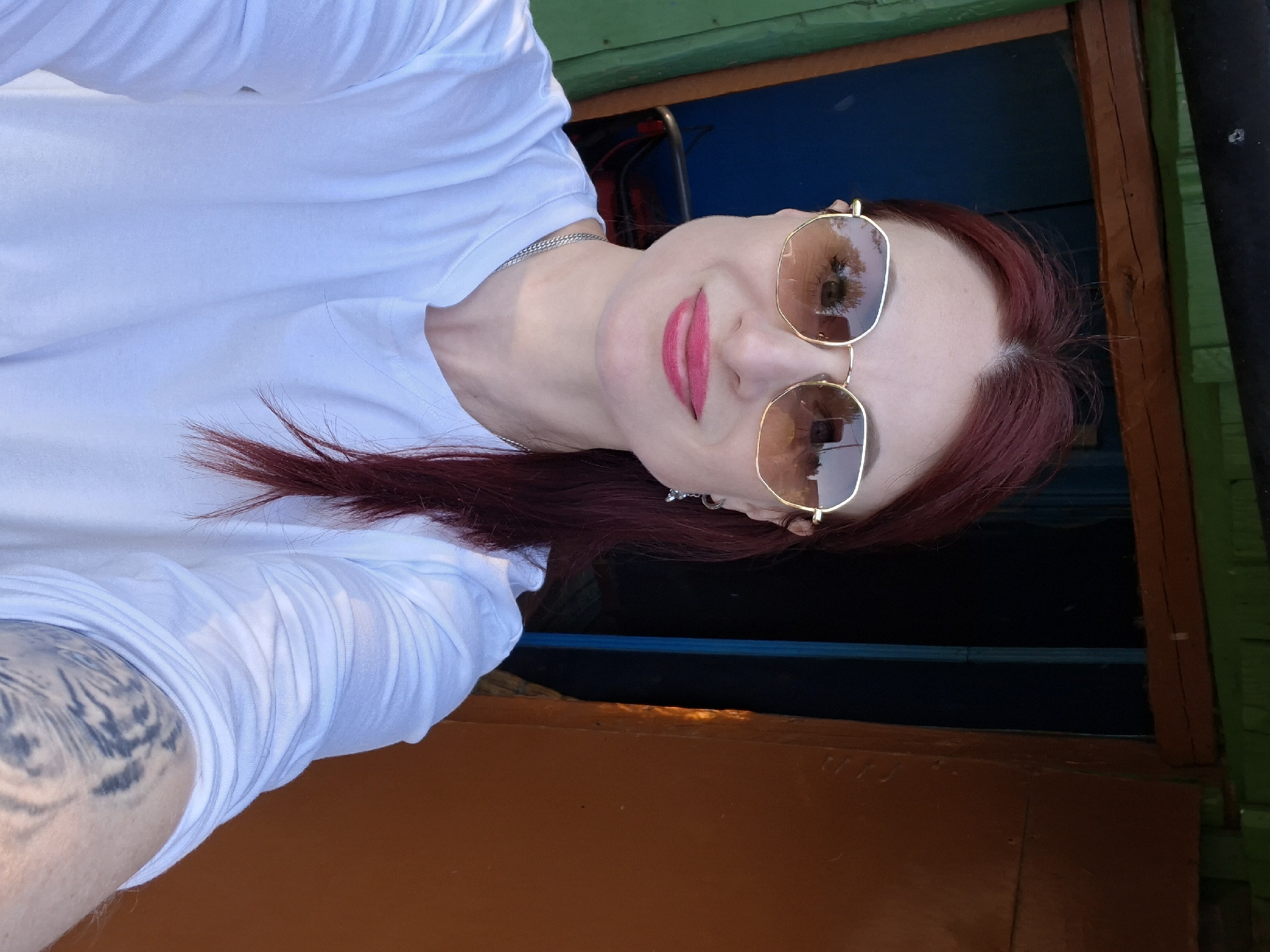 The image is a selfie of a woman with red hair, wearing sunglasses and bright pink lipstick. She is wearing a white t-shirt and has a tattoo on her left arm. The background appears to be a window frame painted in orange, green, and blue. The location is likely Сваткаўскі сельскі Савет, Belarus, based on the information provided. The woman is looking directly at the camera with a slight smile. The image is oriented vertically.