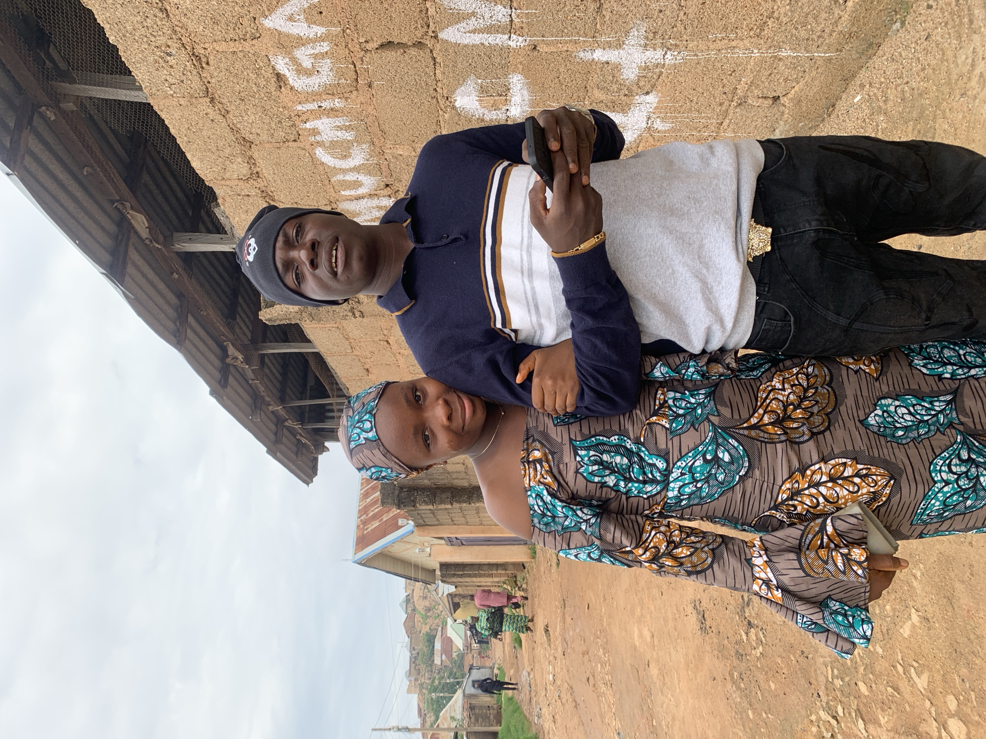 This image depicts two young adults, a man and a woman, standing arm-in-arm on an unpaved street in the city of Jos, Nigeria.
The man, positioned on the right, faces forward with a neutral expression. He wears a dark blue knit beanie featuring a small red and white skull-like logo on the front. His long-sleeved polo shirt is dark blue at the top, transitioning to white, brown, and white horizontal stripes, then a light grey lower section. He wears black jeans with subtle tonal patterns and a prominent gold-colored belt buckle shaped like a Medusa head. A gold-colored bracelet is on his left wrist. He holds a black smartphone horizontally with both hands. His right arm is interlocked with the woman's left arm.
The woman, positioned on the left, smiles faintly and rests her head against the man's right shoulder. She wears an off-the-shoulder dress with a brown background patterned with teal, orange, and white leaf-like motifs. The dress features bell sleeves. A matching patterned headscarf covers her head. She has a thin gold-colored necklace with a small pendant. Her left hand, partially obscured, holds a small, light-colored rectangular object. Her left arm is interlocked with the man's right arm.
The environment features a reddish-brown dirt road in the foreground and extending into the background. Along the road, various single-story buildings with unplastered block walls and corrugated metal roofs are visible; one building presents a light blue gable. A populated hillside with additional dwellings appears in the distant background. Several figures are discernable walking on the road further away. To the immediate right of the individuals is a wall constructed from reddish-brown concrete blocks, bearing white graffiti that includes the word "MUCHIGA" and other faded marks, with white paint drips beneath. Above this wall, a dilapidated structure with a rusty corrugated metal roof, wooden supports, and wire mesh is visible. The sky is overcast and light grey.