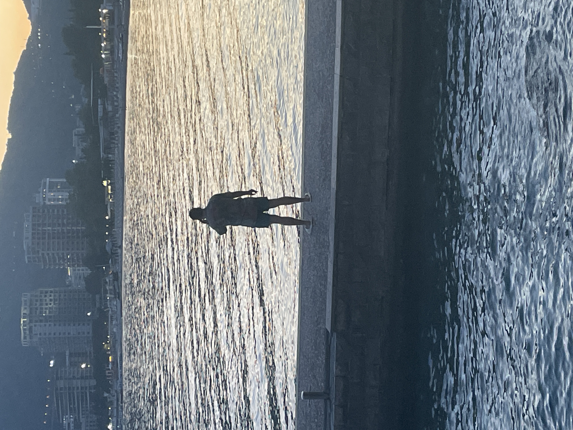 The image depicts a man standing on a concrete pier, silhouetted against a sunlit body of water. In the background, the city of Budva, Montenegro, is visible, with its buildings lining the coastline. The man is positioned centrally in the frame, with his back towards the camera. The water reflects the sunlight, creating a bright, shimmering effect. The pier's edge extends into darker, shaded areas to the right.