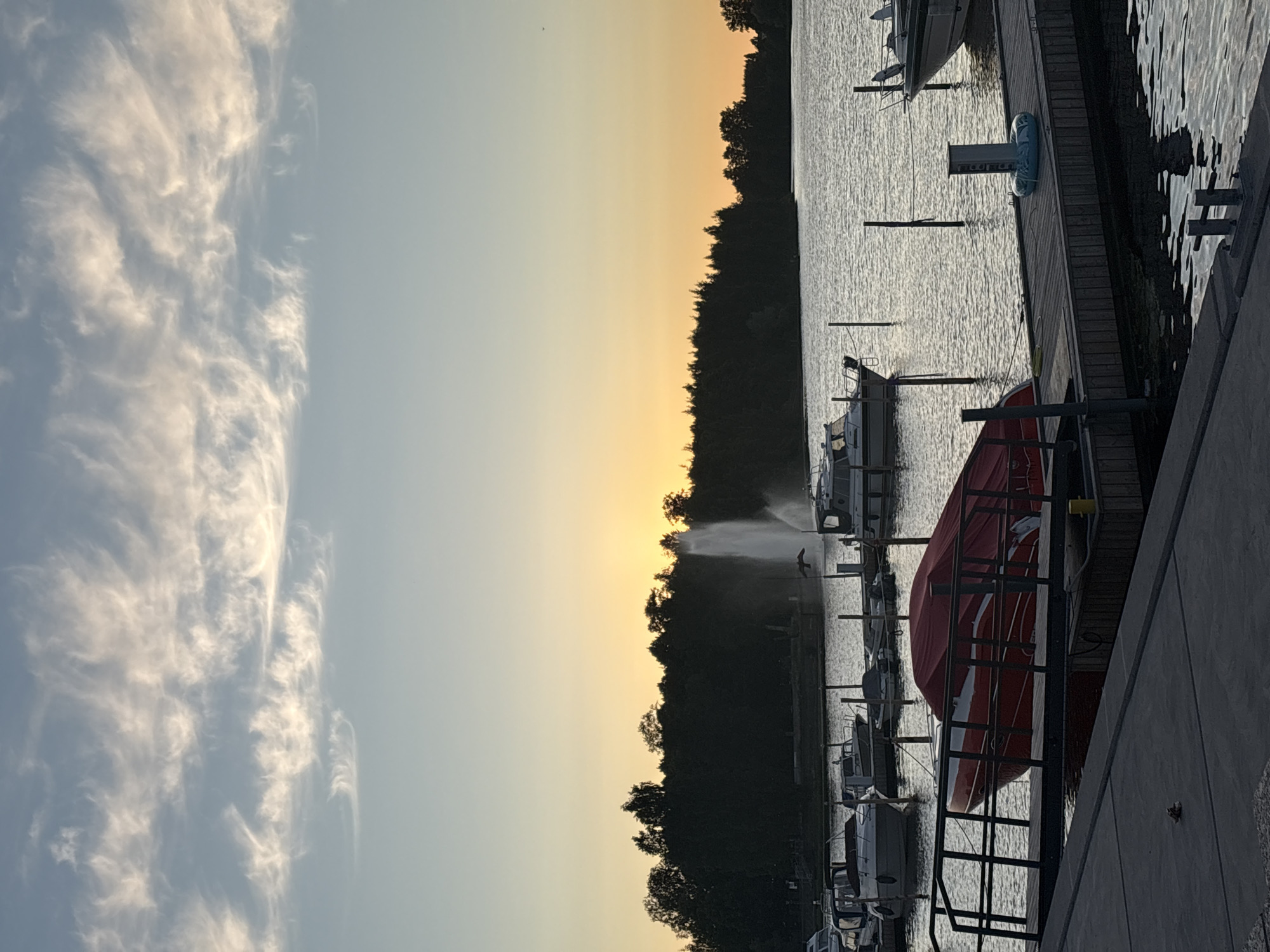 This is a photograph of a marina at sunset in Pori, Finland. The sky is filled with wispy clouds, illuminated by the warm hues of the setting sun. A large fountain sprays water into the air in the middle distance, with a treeline forming the background. Several boats are docked in the marina, including a red boat covered with a tarp in the foreground. The water is calm, reflecting the colors of the sky and the surrounding environment. The scene is peaceful and serene, capturing the beauty of a summer evening in Pori.