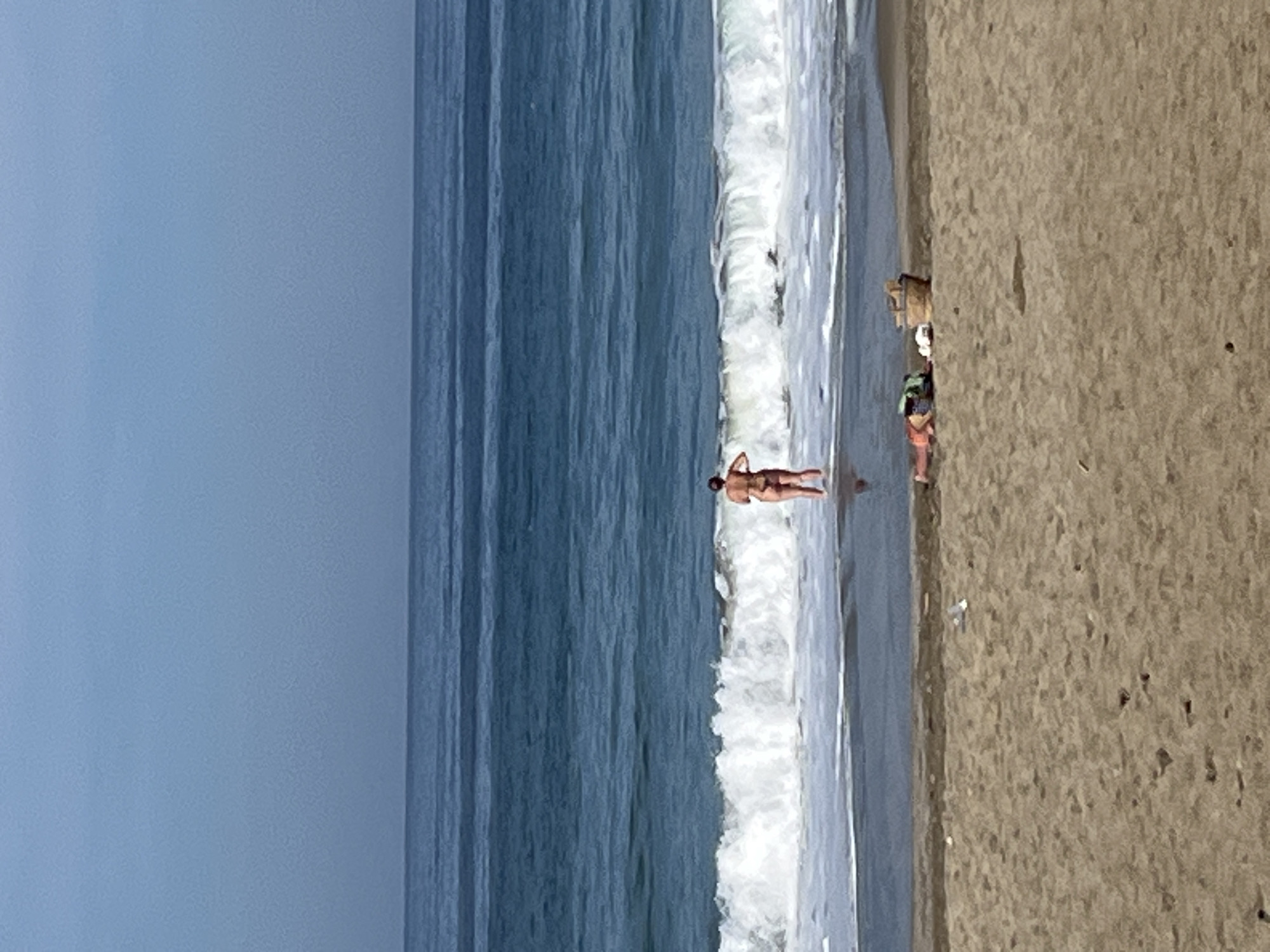 The image shows a beach scene, likely taken in Alanya, Turkey, based on the light and environment. The ocean is a deep blue, with waves breaking along the shoreline. A person stands at the edge of the water, partially submerged by the surf. Further along the beach, other figures are present, including a person with a backpack, and bags. The sand is a beige color. The sky is a hazy blue.