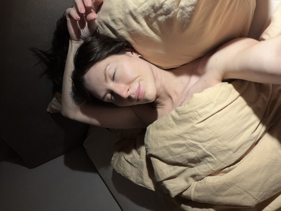 A woman rests in bed, eyes closed, nestled against a beige pillow and wrapped in a matching duvet. Her dark hair contrasts with the pale bedding. A tattoo is visible on her upper arm. The scene is lit with a soft glow, creating shadows on the bed and pillow, adding depth to the composition. The overall impression is one of peaceful slumber and comfort.