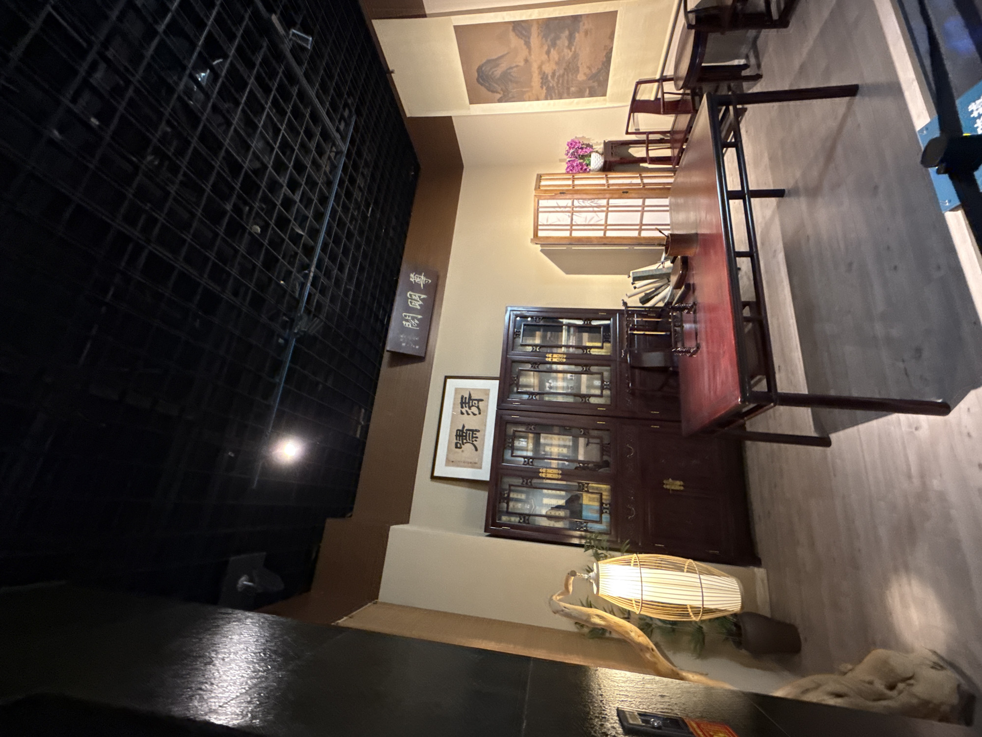 This indoor shot captures a scene in what appears to be an artfully designed space in Yuzhong District, China. Dominating the frame's left side is a large, dark grid-like structure, possibly a window or architectural feature. In the top center is a piece of artwork hanging from the ceiling, a landscape painting.
Further into the space, a wooden table with a dark surface occupies a significant portion of the middle to right side. The scene includes traditional-style furniture, with wooden chairs and what looks like a bookshelf or display cabinet filled with items. The color palette is a mix of dark woods and neutral beige tones. A small illuminated round lamp with a wooden base is positioned near the lower middle of the frame. The objects and arrangement suggests an attempt to create an atmosphere of cultured tradition within this space.