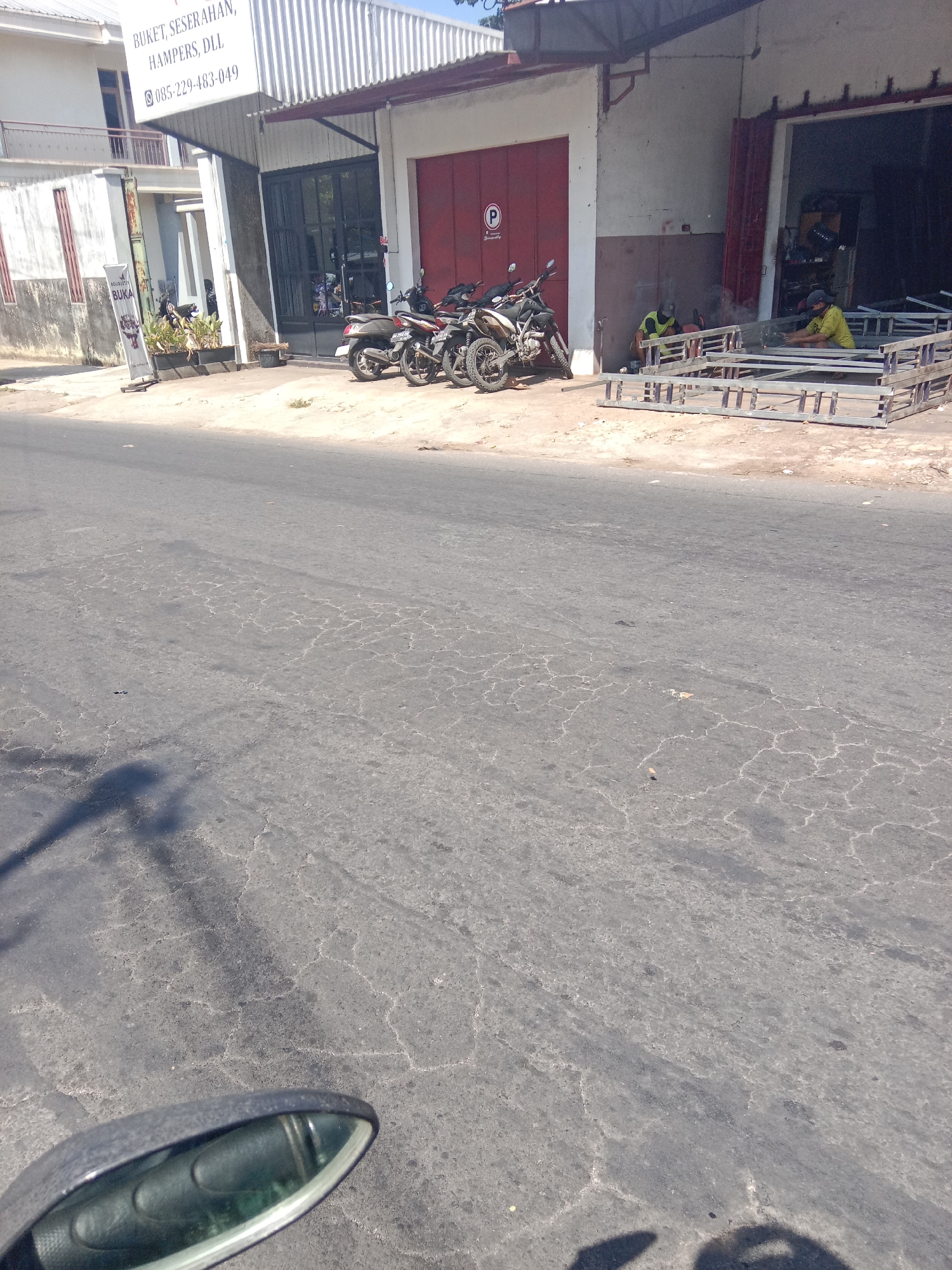 The image depicts a street scene in Dangkel, Indonesia. A building with a white awning and a red garage door is visible on the right. Two men, wearing casual clothing and hats, are working on a metal structure in front of the building. The man on the left is wearing a yellow shirt and appears to be welding, with sparks visible. The man on the right is wearing a blue shirt and is also working on the metal structure. Several motorcycles are parked near the building entrance. A sign on the building has text in Indonesian, including "BUKET, SESERAHAN, HAMPERS, DLL" and a phone number "085-229-483-049." A "P" symbol in a circle is also visible on the red door. The road in front of the building is asphalt and shows signs of wear and tear. A shadow of what appears to be a motorcycle mirror is cast on the road from the perspective of the viewer. The overall environment suggests a commercial or workshop area.