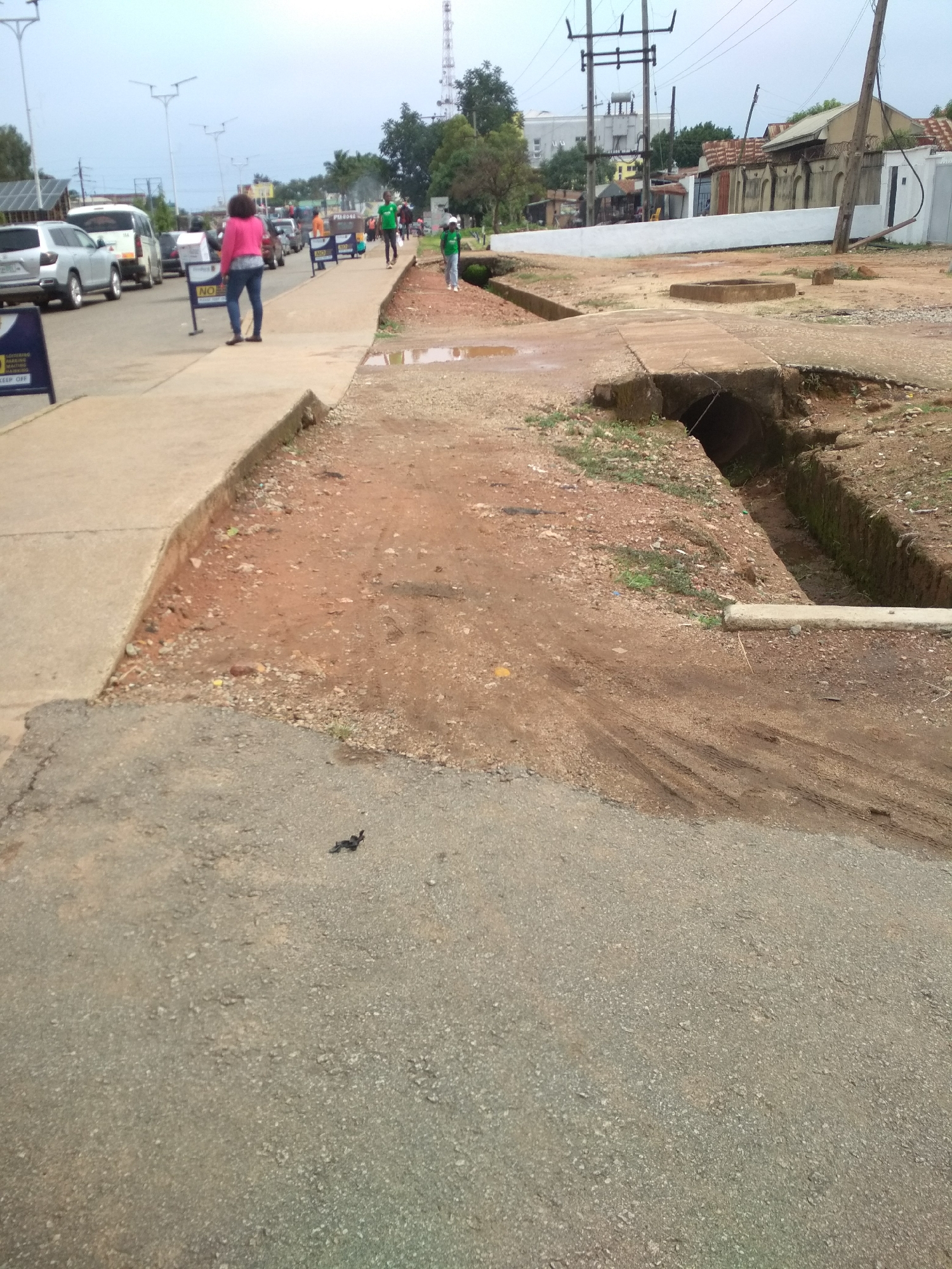 The image depicts a daytime street scene in what appears to be an urban or semi-urban area, consistent with Jos, Nigeria. The sky is overcast, and the lighting is soft.
In the foreground, a paved road surface with small gravel leads to a raised concrete sidewalk. Beyond the sidewalk, an unpaved, reddish-brown dirt area with some sparse vegetation extends, revealing significant infrastructure deficiencies. A large, deep ditch or open channel runs parallel to the sidewalk, with exposed concrete culverts and eroded banks, suggesting a drainage system or a construction site. Puddles of water are visible in the dirt area, indicating recent rainfall or poor drainage. Tire tracks are visible in the muddy ground near the ditch.
Several people are present. On the sidewalk, a woman in a pink top and blue jeans walks away from the viewer. Further down the street, other pedestrians, including individuals in green shirts, are visible. Cars, including a silver SUV and a white van, are moving or parked on the road, indicating traffic. A tricycle (keke napep) is also seen in the distance.
Various urban objects are scattered throughout the scene. Prominent utility poles with power lines and a tall antenna or cell tower are visible against the skyline. In the background, buildings with varied architecture, some with rusty corrugated roofs and a long white wall, line the street.
Visible text on signs includes "NO PARKING" and partially obscured text that reads "CONTAINING PARKING WORKING" and "KEEP OFF". The overall impression is one of mixed development, with functional roadways and sidewalks adjacent to significantly underdeveloped or damaged unpaved areas and open ditches.