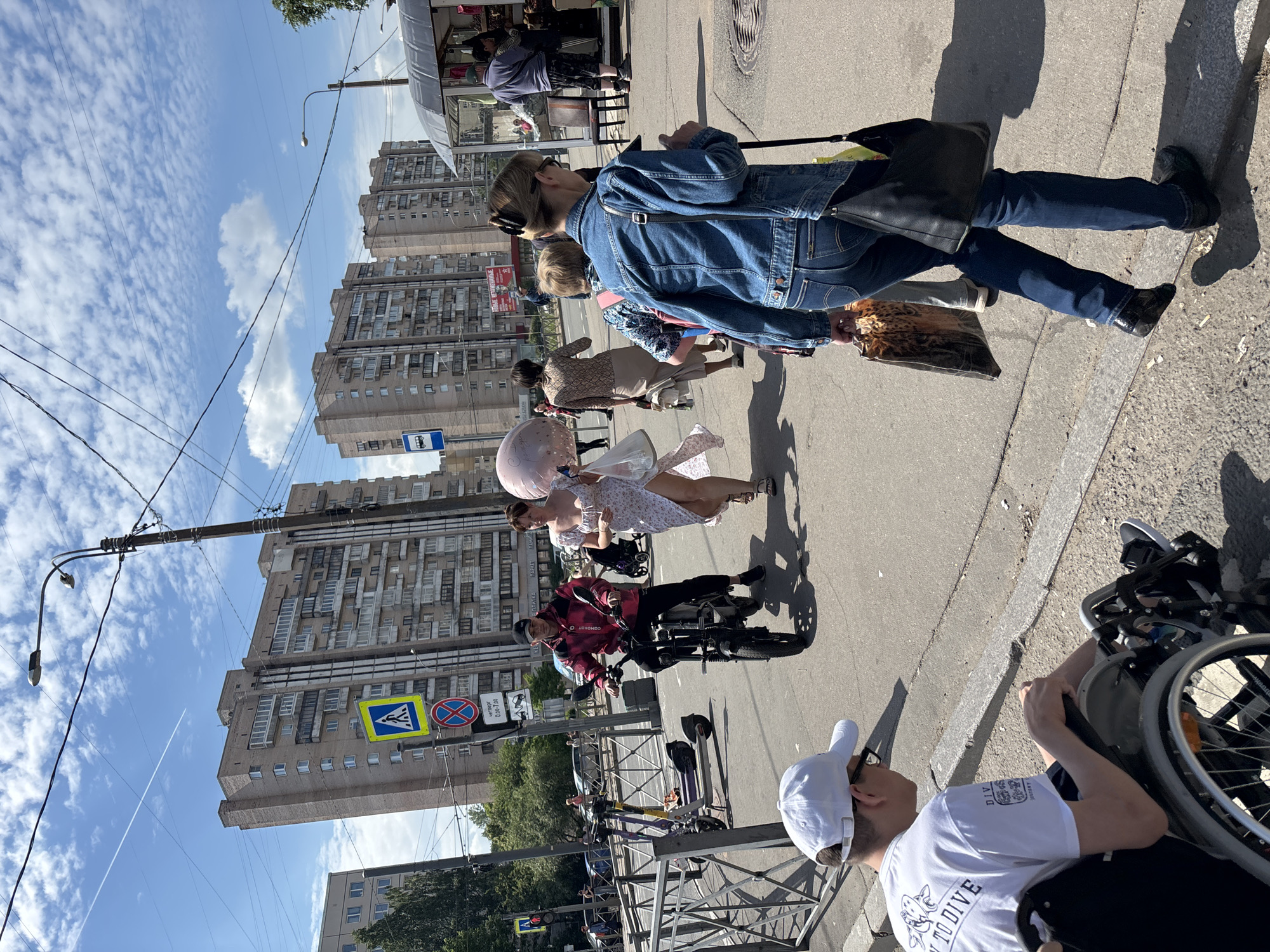 The image captures a vibrant street scene on a clear, sunny day in an urban setting, characteristic of Russia or Eastern Europe. The sky is bright blue with scattered white clouds, and strong sunlight casts distinct shadows.
In the foreground, a person wearing a white baseball cap and a white t-shirt with "DIVING TO DIVE" visible text is seated in a wheelchair on the sidewalk. To their left, another person on a small electric scooter or motorcycle, wearing a red plaid shirt (with "COMPACT" text partially visible) and black pants, is positioned in the street.
Further back, several pedestrians are visible. A woman in a light patterned dress holds a large pink balloon, while another woman walks beside her in a brown patterned outfit. To their right, a woman dressed in a denim jacket and jeans walks away, carrying a black shoulder bag and a leopard-print tote bag. In the background, other individuals are seen near a street vendor's stall with an awning.
The street is lined with tall, multi-story residential buildings featuring numerous windows and balconies, common to post-Soviet architecture. Street infrastructure includes power poles, overhead wires, and streetlights. Several street signs are visible: a standard blue bus stop sign, a yellow diamond sign indicating a pedestrian crossing, and a red circular sign for "no stopping/parking." A smaller sign below the bus stop sign contains Cyrillic text, likely detailing bus routes or schedules. People are engaged in everyday activities like walking and commuting.