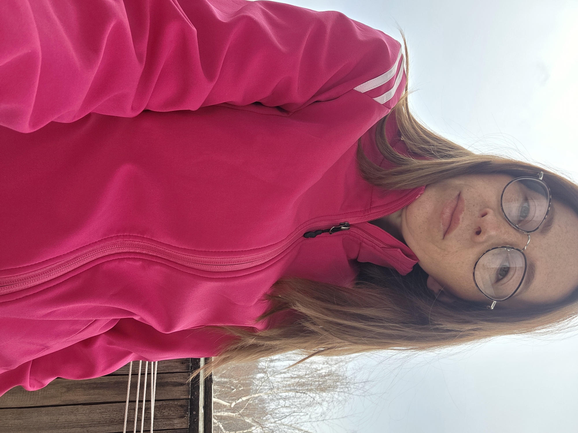 A woman wearing a pink jacket and round glasses is looking down at the camera. The picture was taken outdoors. The bottom of the frame shows a wooden platform or railing with snow-covered ground visible beneath. Above the woman is a pale sky. Given the context of Chaikovsky, Russia, it is likely a cold climate.