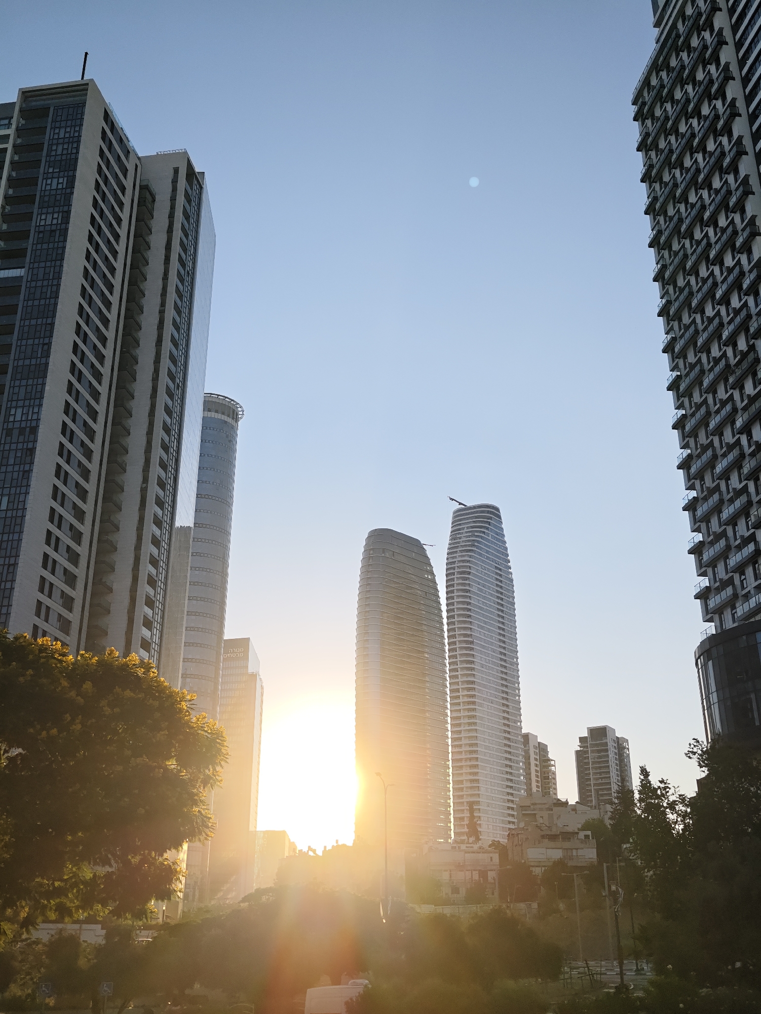 The image captures a street view in Givatayim, Israel, during a sunset. Tall, modern buildings dominate the skyline, with their reflective surfaces catching the golden light. The sun, a bright orb, is positioned low in the sky, casting long rays that create a lens flare effect across the scene. Some of the buildings are sleek and cylindrical, while others are more angular and rectangular, showcasing a variety of contemporary architectural styles. Greenery, including trees and bushes, is visible in the foreground and interspersed between the buildings, adding a touch of nature to the urban landscape. The sky is a clear, pale blue, with a faint full moon visible in the upper left quadrant of the image. The overall impression is one of a vibrant city bathed in the warm glow of the setting sun.