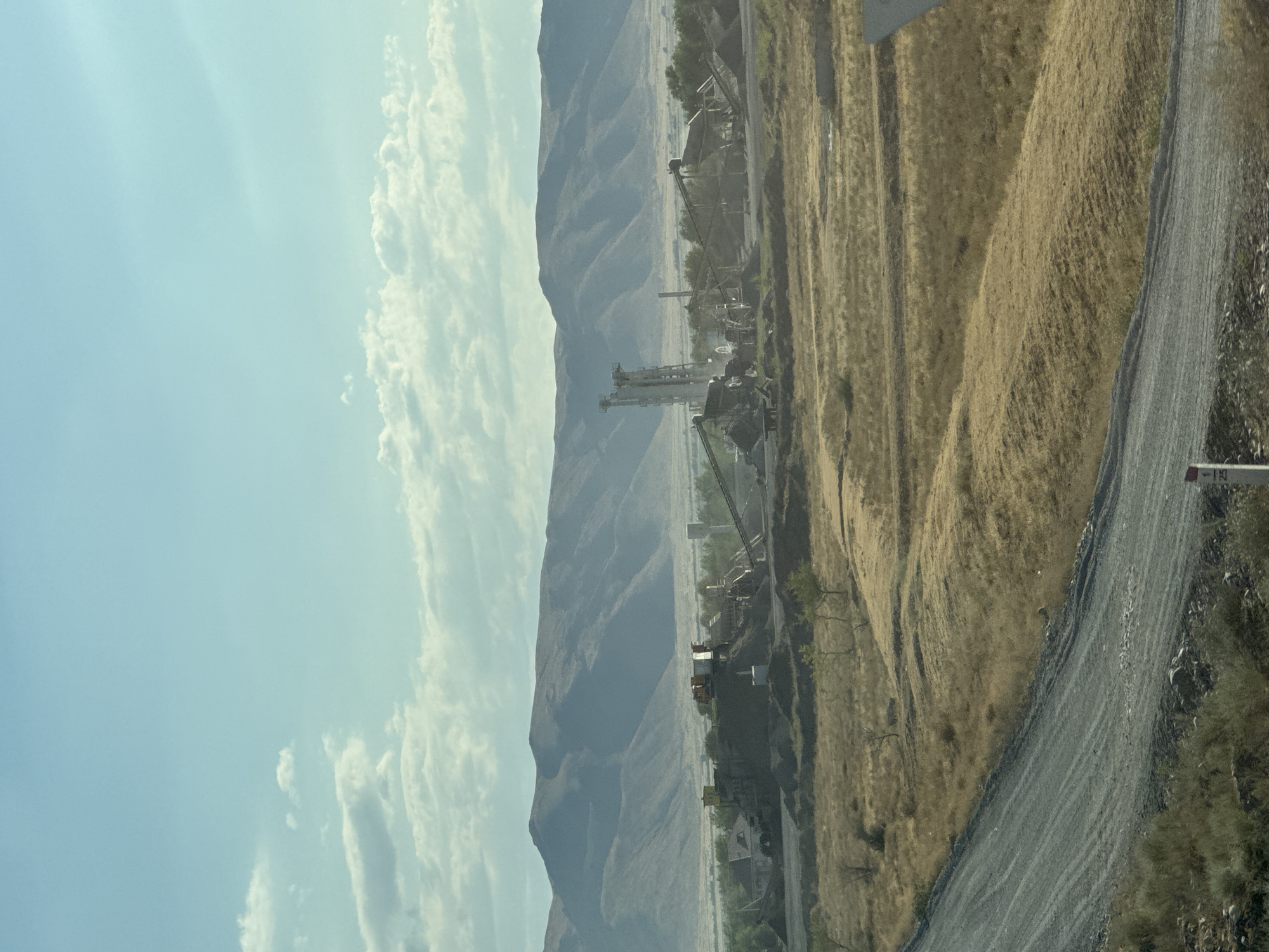 The image depicts a wide-angle view of an industrial facility situated in a rural, arid landscape. In the foreground, a gray, unpaved road winds from the lower left corner upwards and to the right. The surrounding terrain is characterized by dry, golden-brown grasses and sparse vegetation, with some rolling hills.
Dominating the mid-ground is a complex of industrial structures, likely a processing plant or quarry. Tall, tower-like machinery, conveyor belts, piles of dark material (possibly gravel or ore), and smaller buildings are visible. The structures appear functional, suggesting ongoing operations.
In the background, a range of muted, layered mountains stretches across the horizon. The sky is a pale blue with scattered white, wispy clouds, indicating fair weather. The overall impression is one of industry integrated into a natural, somewhat stark environment. The location is identified as Üşaral, Kazakhstan.
