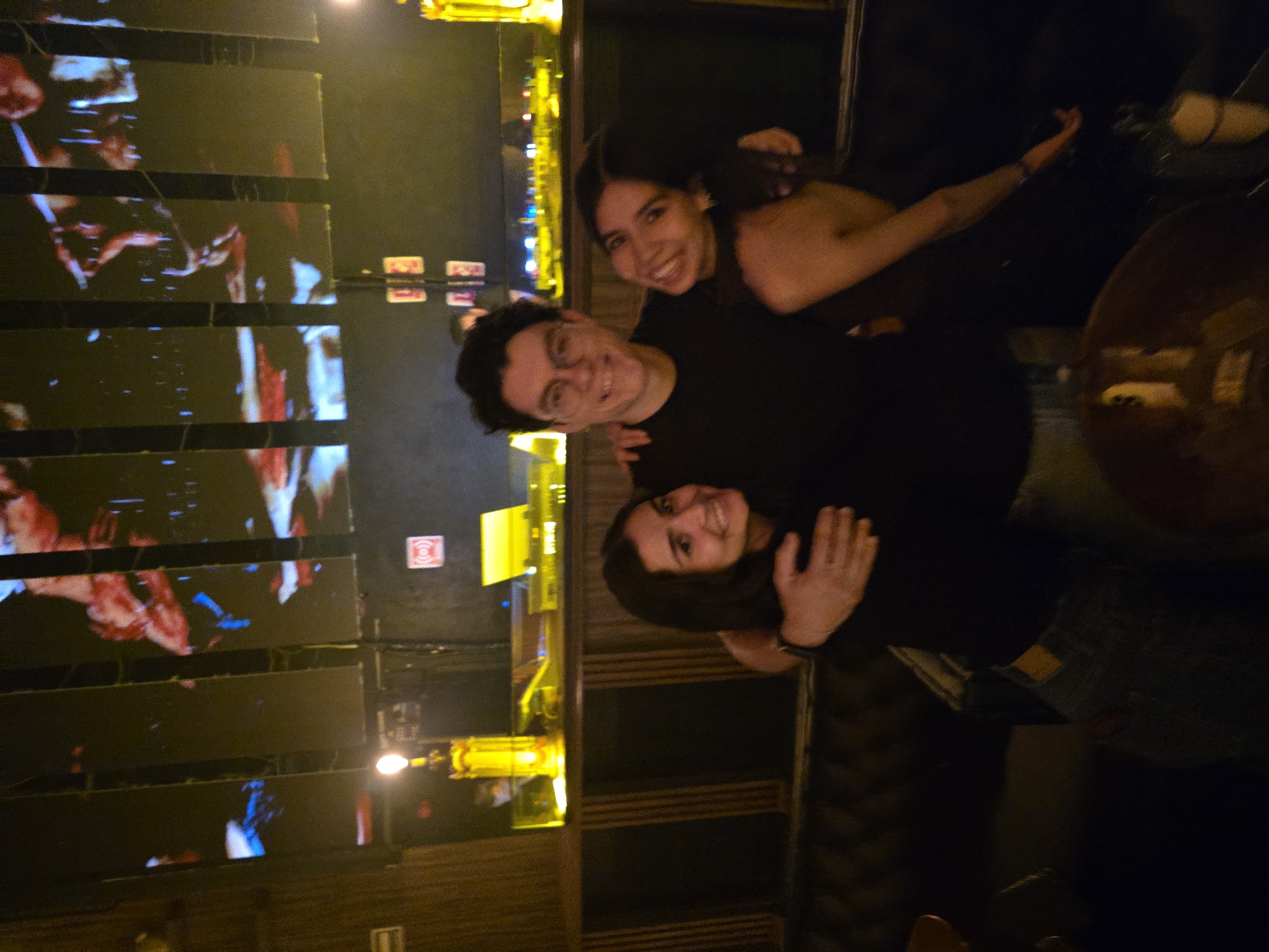 Three individuals are grouped together in what appears to be an indoor venue with low lighting. The setting is a bar or entertainment space, indicated by wall-mounted displays and accent lighting. A male with dark hair and glasses, wearing a dark shirt, is positioned in the middle, with two women on either side of him. One of the women is partially visible, with dark hair and exposed shoulders. The other woman is closer to the camera and has her hand raised near her face. The group is tightly knit, suggesting a friendly or familiar relationship. The decor features a combination of dark, neutral tones and vibrant lighting fixtures. In the background, segmented displays exhibit abstract patterns or images, adding to the atmospheric ambiance. The photograph was taken in Puebla, Mexico.