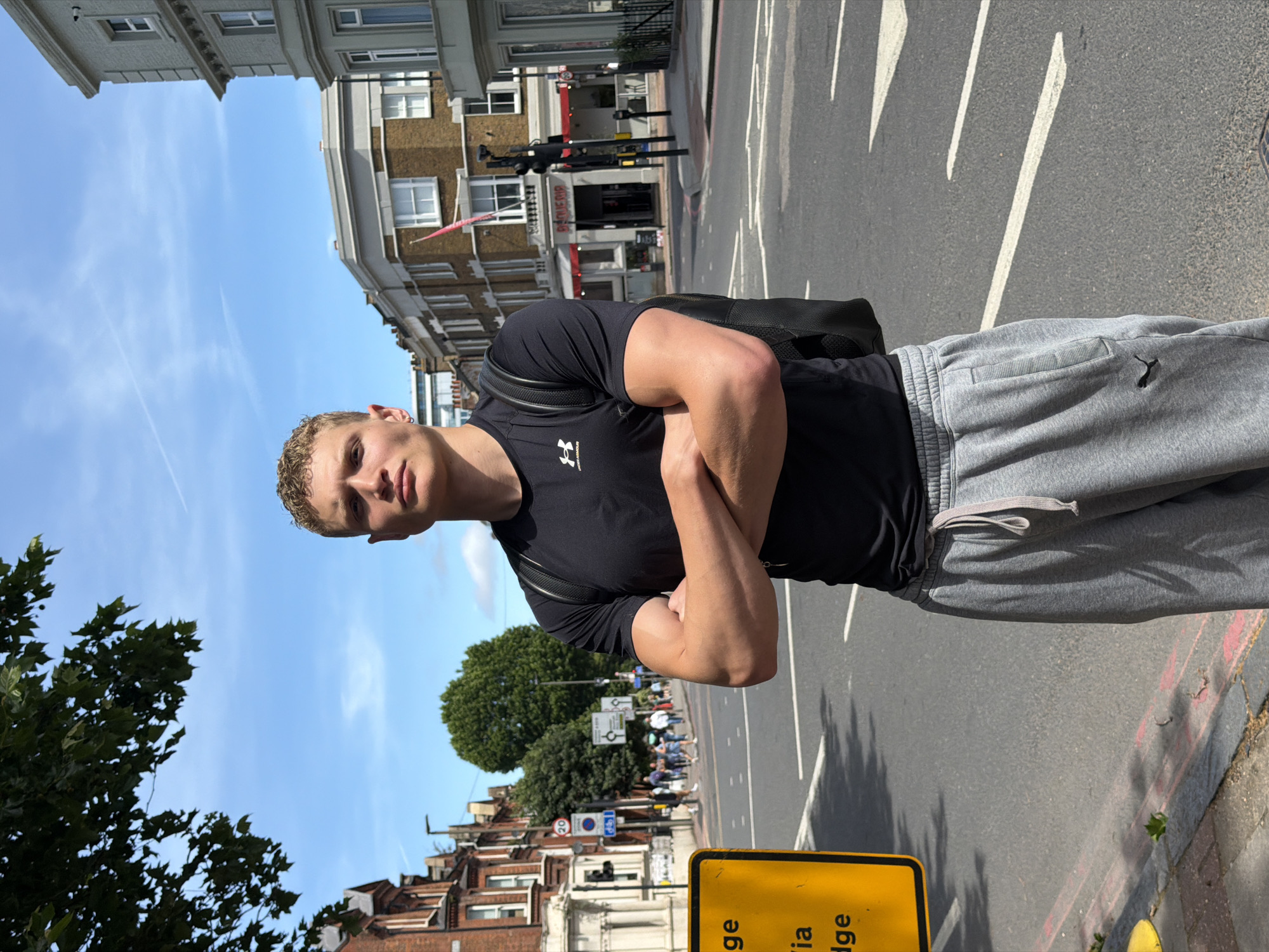 A young, muscular man with short blonde hair stands on a city street with his arms crossed. He is wearing a black t-shirt with an Under Armour logo and grey sweatpants with a Puma logo. He also has a black backpack on. The street is paved with asphalt and has white road markings. There are buildings in the background, including red brick houses and a building with a light-colored facade. A yellow street sign with black text is visible on the left side of the image. The sky is blue with some wispy clouds. The man appears to be in London, United Kingdom, as indicated by the presence of a red double-decker bus, a black cab, and signage consistent with UK road regulations, such as the "20" speed limit sign.