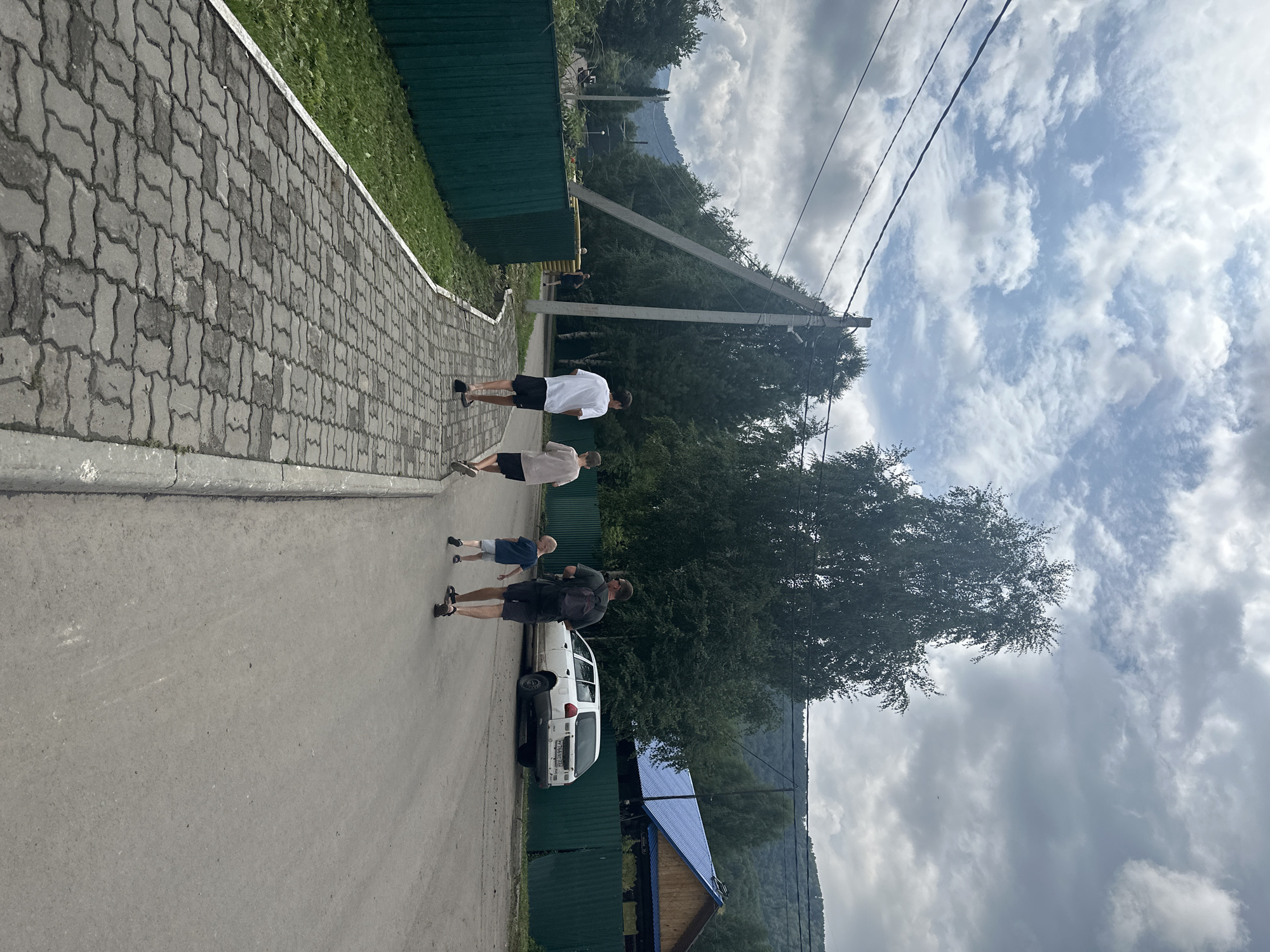 The image depicts a family walking on a paved pathway next to a road. A man, two boys, and a young child are walking away from the camera. The man is wearing a dark t-shirt and shorts, with a backpack. The boys are wearing t-shirts and shorts. The young child is wearing a blue t-shirt and shorts. A white car is parked on the side of the road. A green fence runs along the left side of the pathway. In the background, there are trees, a house with a blue roof, and a hill. The sky is cloudy. The location appears to be a rural or semi-rural area in Danilovka, Altai Krai, Russia, as indicated by the presence of power lines, a house with traditional Russian architectural elements, and the natural landscape.