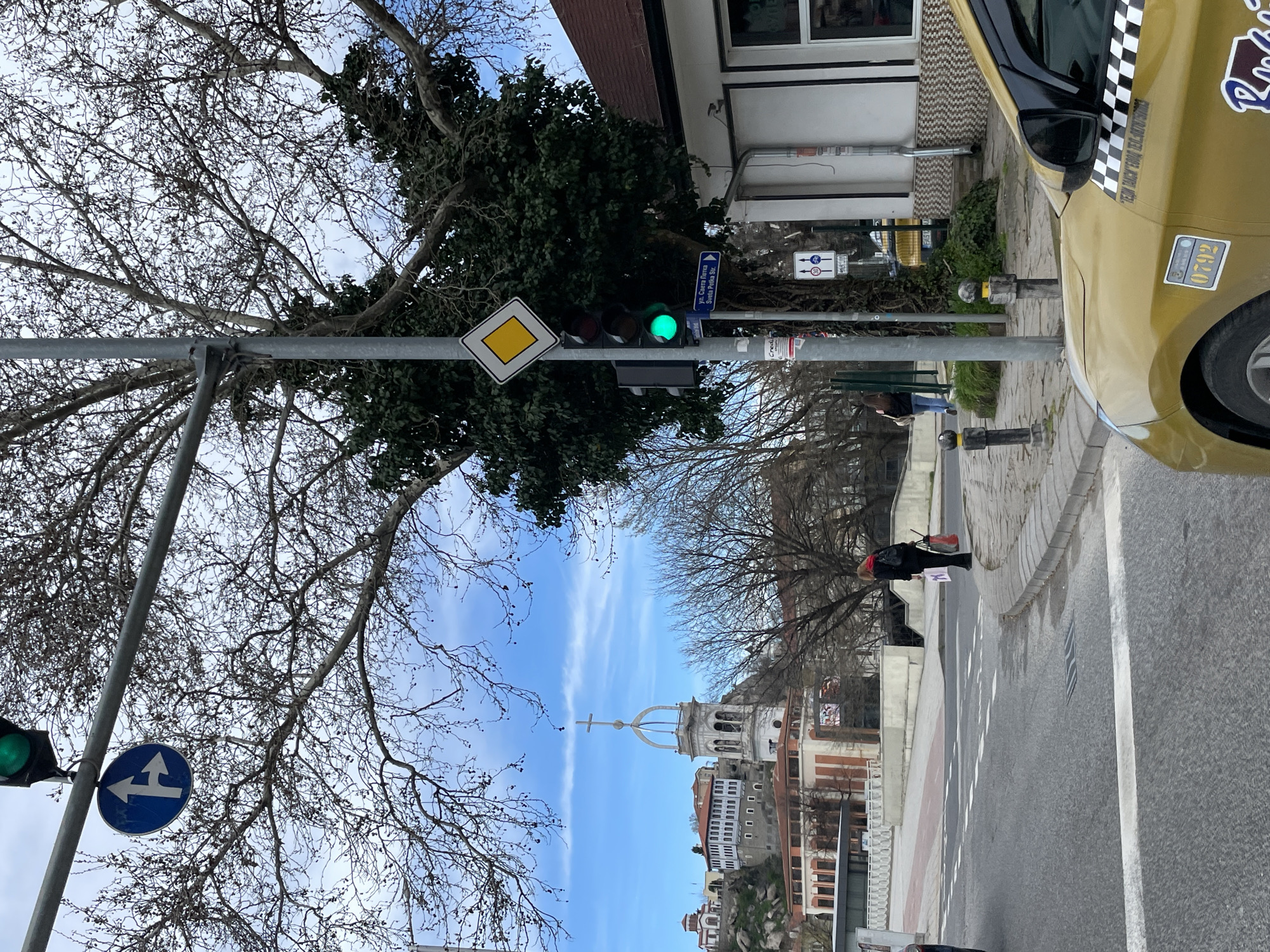 The image depicts a street scene. A traffic light is visible, showing a green light. A yellow diamond-shaped sign is mounted on the same pole as the traffic light, along with another blue circular sign. A street sign reading "ул. Света Петка Sveta Petka Str." is also attached to the pole. A tree with bare branches is on the left side of the image, and another tree with green foliage is near the traffic light. A building with a white facade is in the background. A taxi cab is partially visible on the right side of the frame, showing its license plate "0792" and the company name "ТЕДИ ТАКСИ". In the distance, there is a building with a prominent spire. The scene is set in Plovdiv, Bulgaria, specifically on Sveta Petka Street.