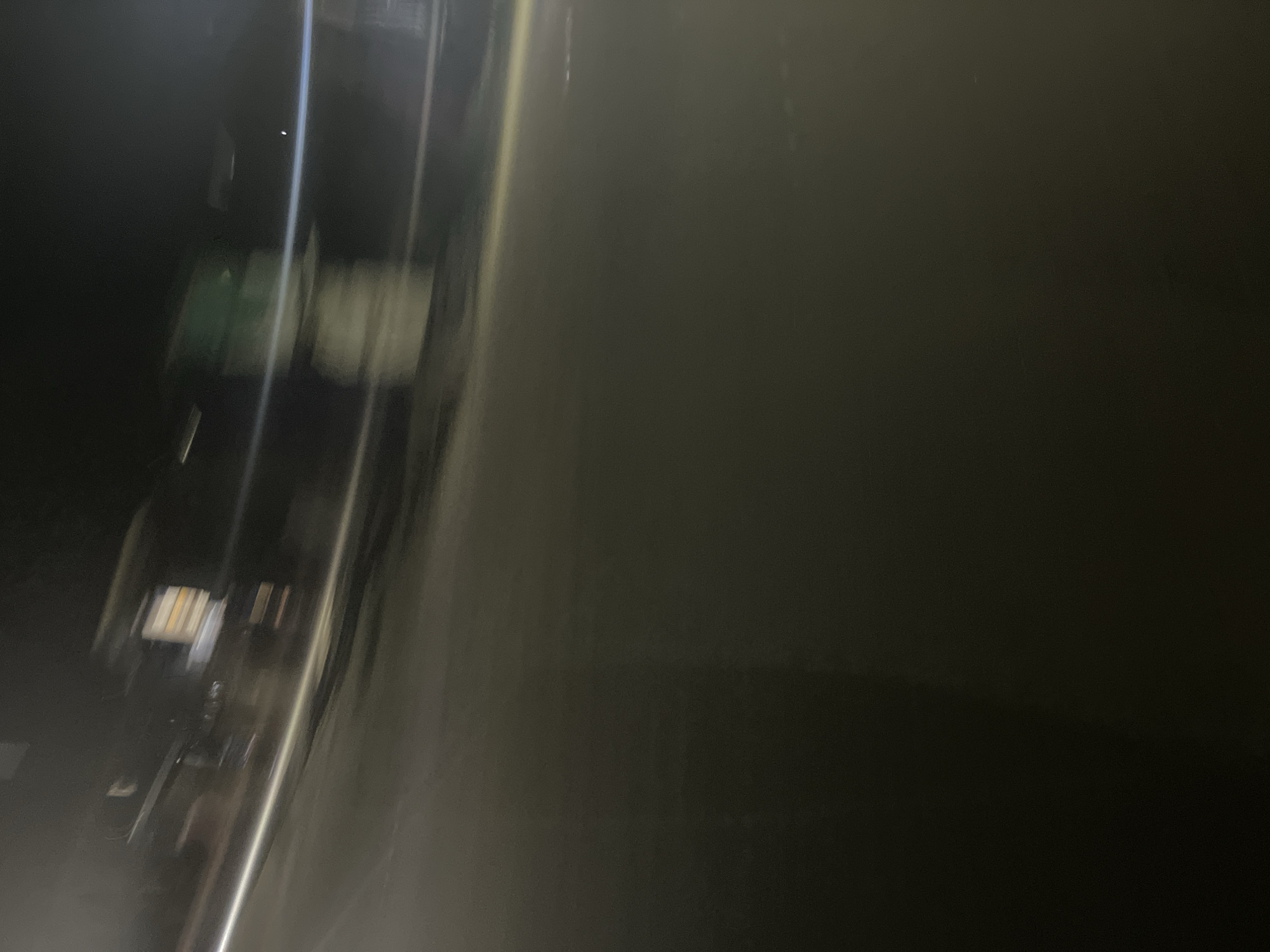 The image, taken at night in an urban environment, appears to be a motion-blurred photograph of a street. In the upper portion of the image, there are streaks of light and blurred shapes that suggest movement and the presence of buildings or structures. One building shows illuminated windows. A horizontal band of light, possibly from streetlights or vehicle headlights, cuts across the scene. The foreground is dark and appears to be a paved surface, likely a road. The overall impression is one of a nighttime streetscape experienced during travel, perhaps from a moving vehicle.
