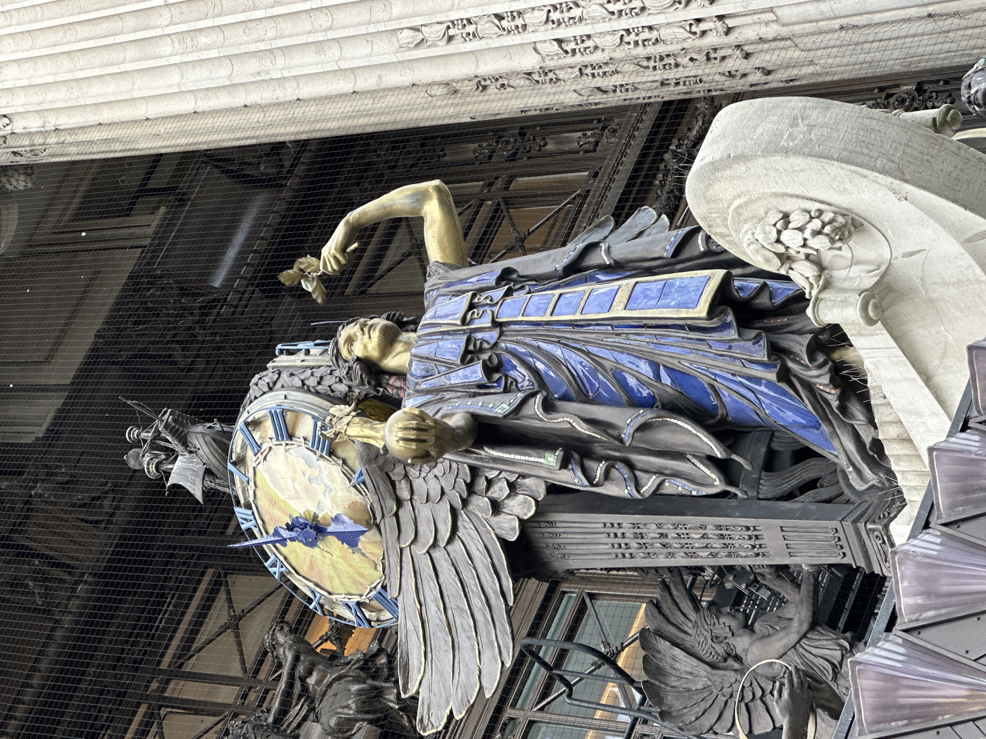 The image depicts a portion of the Selfridges Clock in London, United Kingdom. It features a golden statue of a woman lying on her side, draped in blue and black fabrics. She holds a small object in her raised right hand. A large, ornate clock face is visible beneath her, with blue Roman numerals and arrow-shaped hands. Winged sculptures support the clock structure, and a filigree roof and safety netting are visible in the background. The architectural elements suggest the exterior of a building.