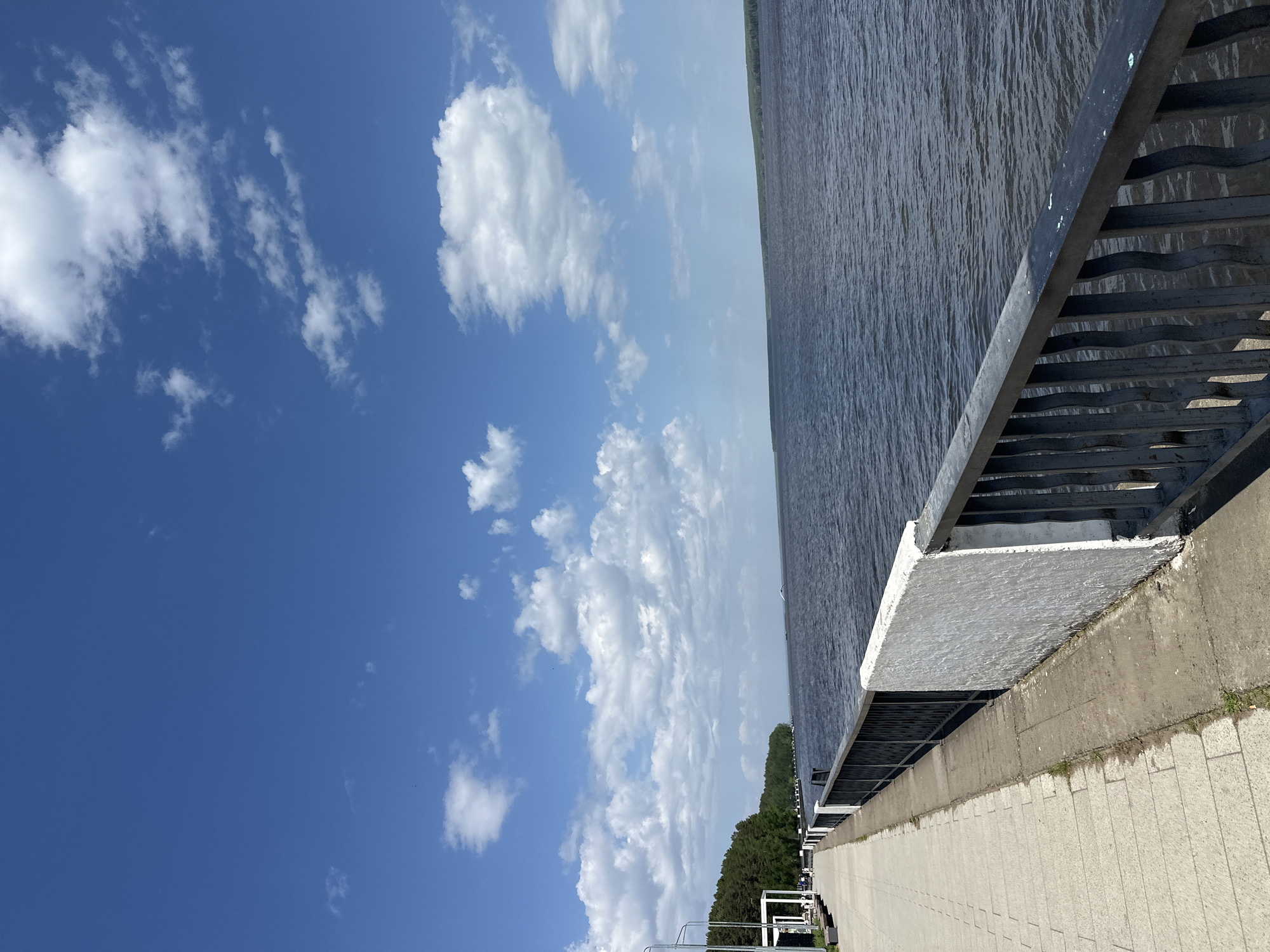 View of the Kama River from the embankment in Chaikovsky, Russia. The sky is blue with scattered cumulus clouds. To the right, a metal railing lines the riverbank, which is paved with stone. A line of trees can be seen in the distance along the opposite shore.