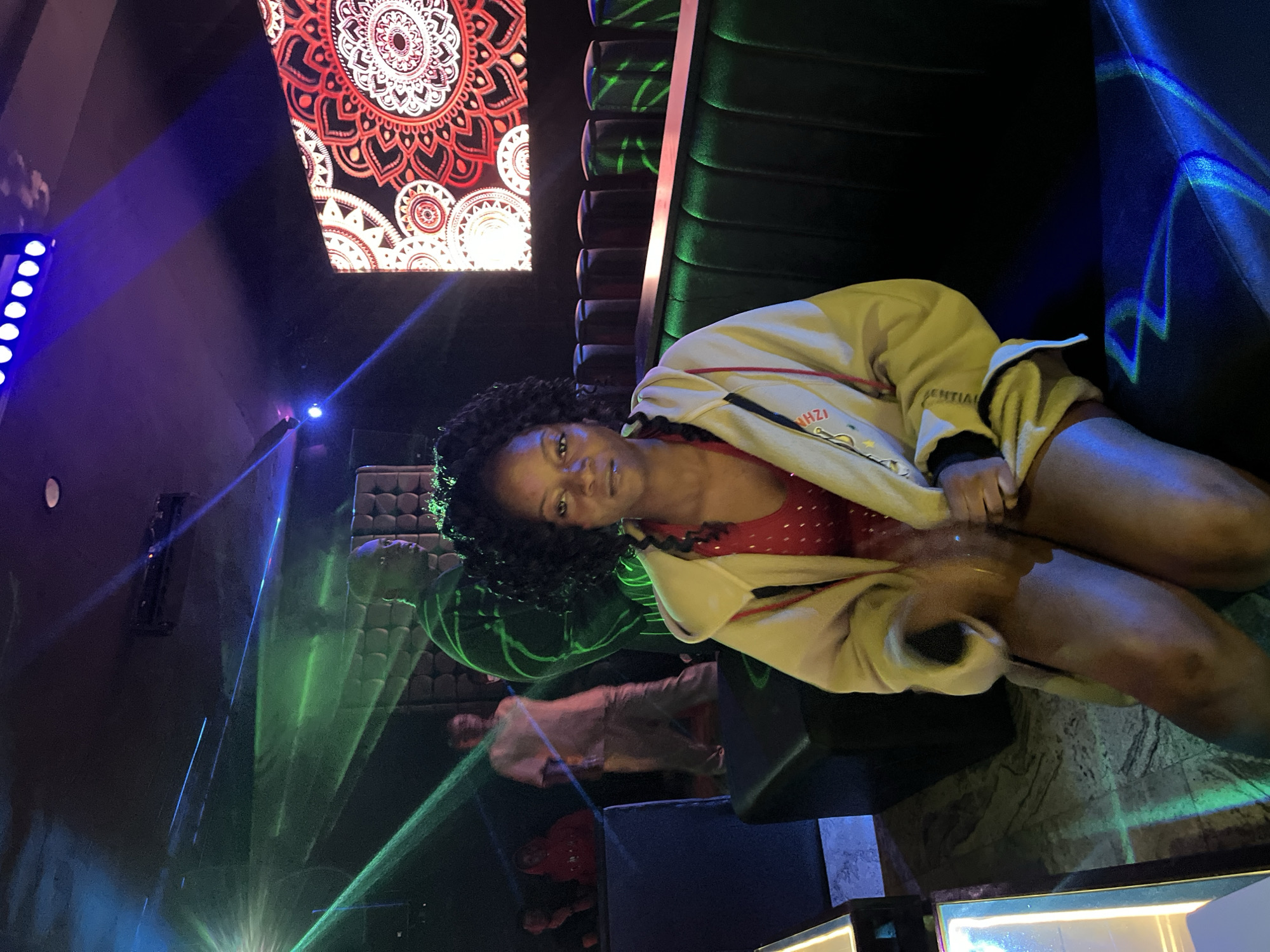 This image depicts an indoor nightlife setting, specifically a lounge or club, located in the city of Howayi, Nigeria.
In the foreground, a dark-skinned female individual is seated on a dark, possibly green, upholstered bench or booth. She has dark, curly hair and is wearing a red top with visible texture or pattern, layered under an open, light-colored (beige or cream) hooded jacket. Her hands are in her lap, slightly blurred, indicating motion or a longer exposure. She is looking directly at the viewer. Her bare legs are visible.
The environment is dimly lit with various artificial light sources. Green and blue laser beams crisscross the space, illuminating dust particles in the air and casting patterns on surfaces and individuals. Overhead, multiple blue LED lights are visible, casting light downwards.
In the mid-ground and background, several other individuals are present, most of whom are out of focus or partially obscured. One dark-skinned male figure stands in the mid-left, partially illuminated by the green laser light. Another male figure in the further background is also visible. Further left, two more figures are seated or standing, one wearing a red hoodie.
On the right side of the frame, a large digital display screen projects a vibrant red and black intricate, mandala-style pattern. The seating area features dark, upholstered booths with visible backrests. The floor appears to be a dark, possibly marbled or patterned, surface, reflecting some of the laser lights. A dark, cuboid object, possibly a speaker or table base, is situated on the floor to the left of the main subject.