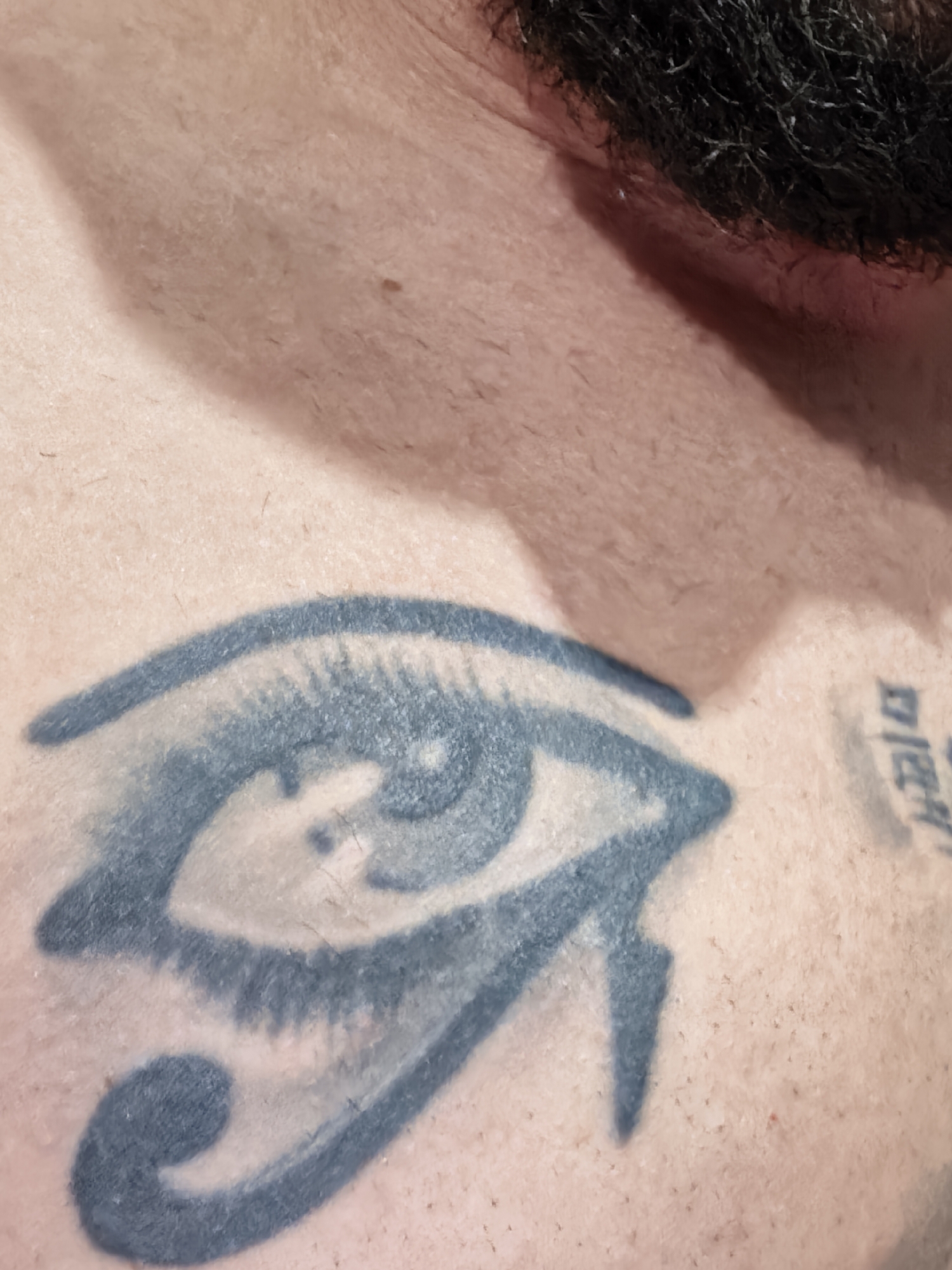 The image shows a close-up of a person's upper chest and neck area. Visible are two tattoos on the person's skin. One tattoo appears to be the Eye of Horus, rendered in a blue-gray ink. Slightly off to the right and partially out of frame, is another tattoo featuring indiscernible symbols. The person has light skin, with stubble visible, and a patch of dark, coarse hair is present at the top of the frame, presumably part of a beard or sideburn. The location is not identifiable in the image.